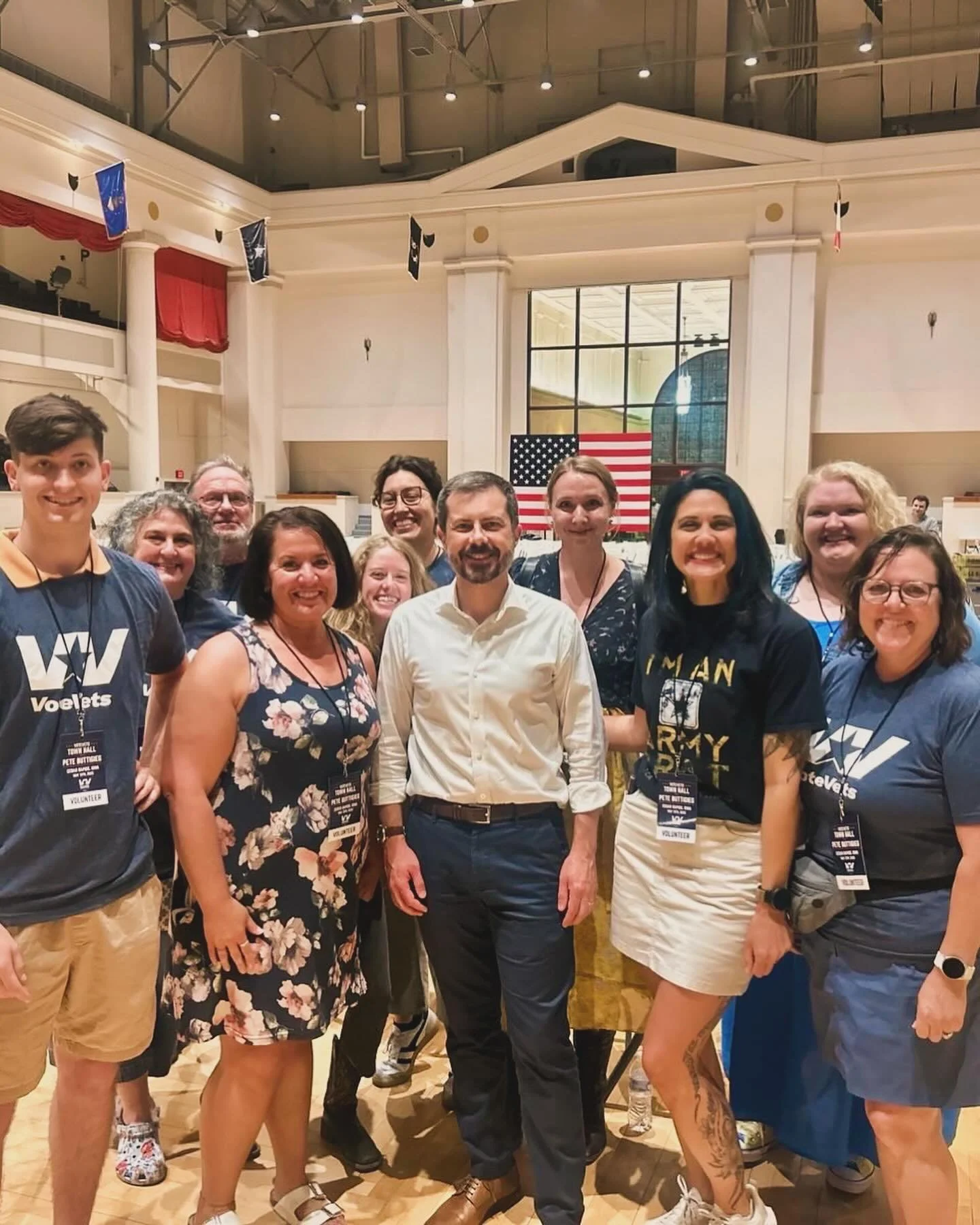 Group of people standing in an indoor hall with an American flag in the background.