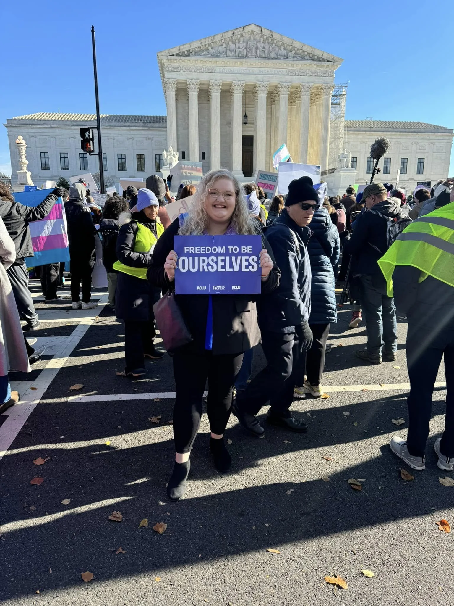 A woman smiling and holding a sign that says "FREEDOM TO BE OURSELVES" at a protest or rally in front of the U.S. Supreme Court building, with a crowd of people around her holding signs and flags.