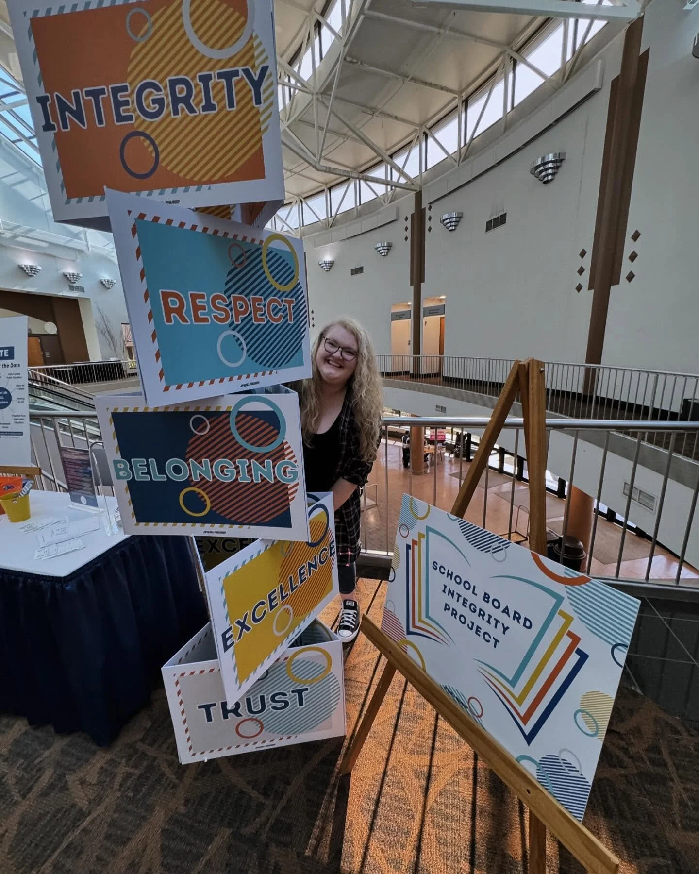 A woman with curly blonde hair and glasses is smiling behind a display of colorful signs that read: 'INTEGRITY', 'RESPECT', 'BELONGING', 'EXCELLENCE', and 'TRUST'. Next to her is a large sign board that says 'School Board Integrity Project'.