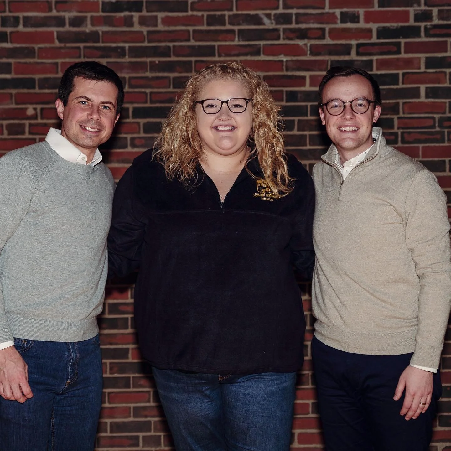 Three people standing together in front of a brick wall, smiling at the camera. The person in the middle is a woman with curly blonde hair and glasses, wearing a black zip-up jacket. The two men on either side of her have short dark hair, are wearing
