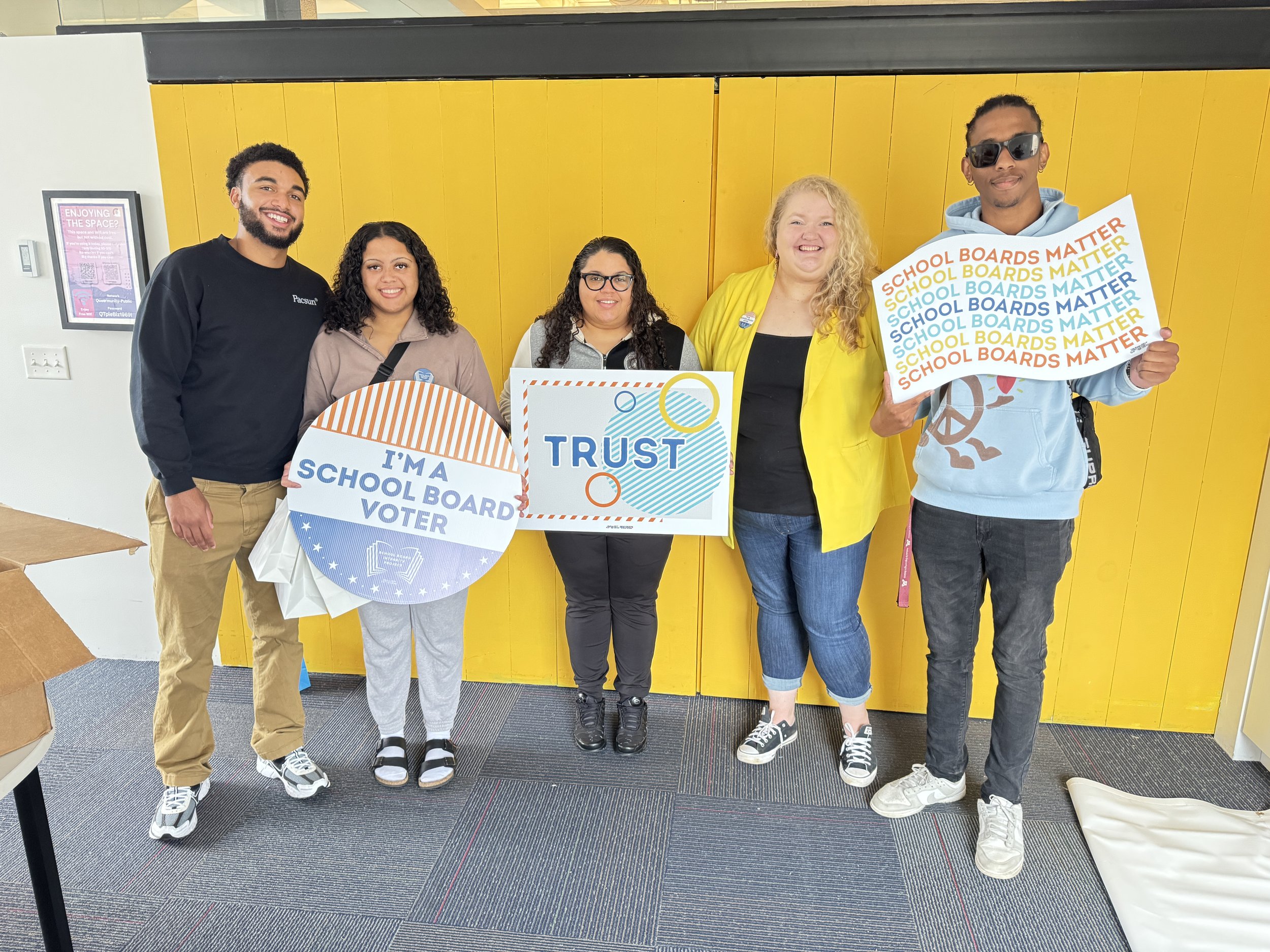 Group of five diverse people standing in front of a yellow wall, holding signs supporting school board voting and trust, with some smiling and wearing casual clothes.