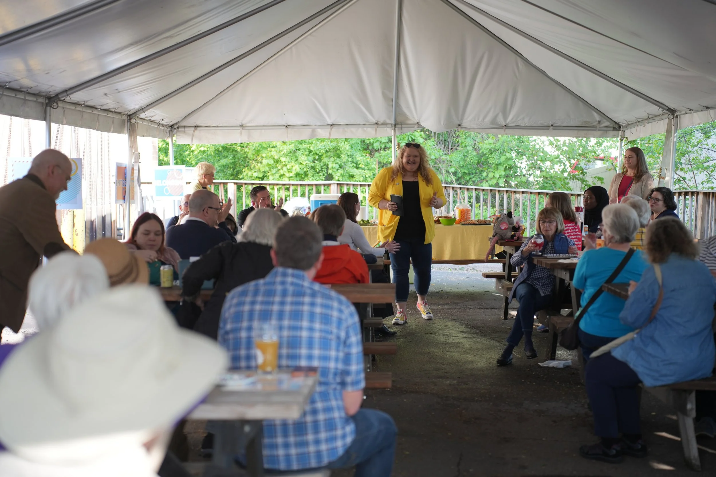 People gathered under a large outdoor tent for a social event, some seated at picnic tables and others standing, with a woman in a yellow jacket standing and speaking to the group.