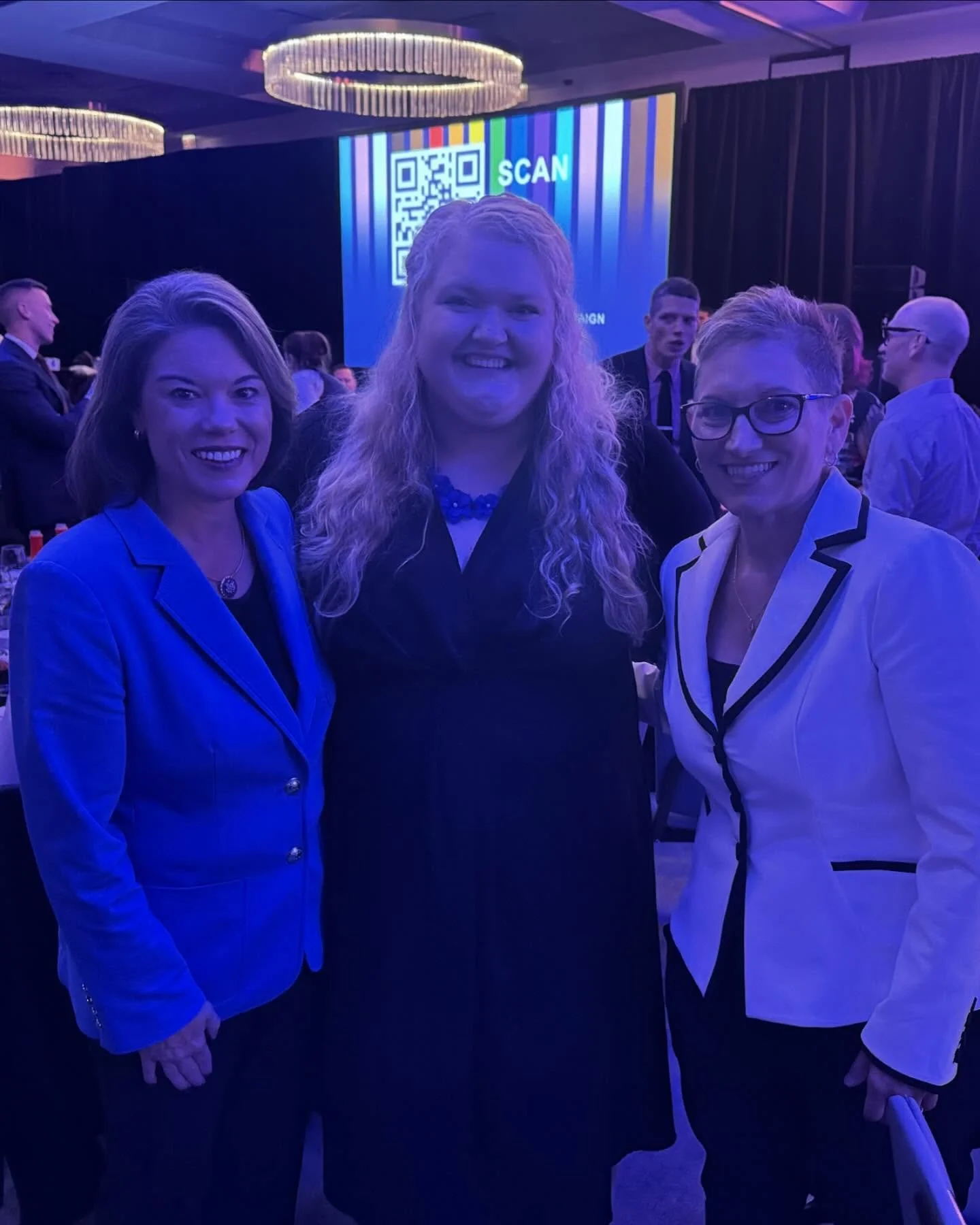 Three women in business attire smiling at a conference or event, with a large screen behind them displaying a QR code and the word 'SCAN' in colorful text.