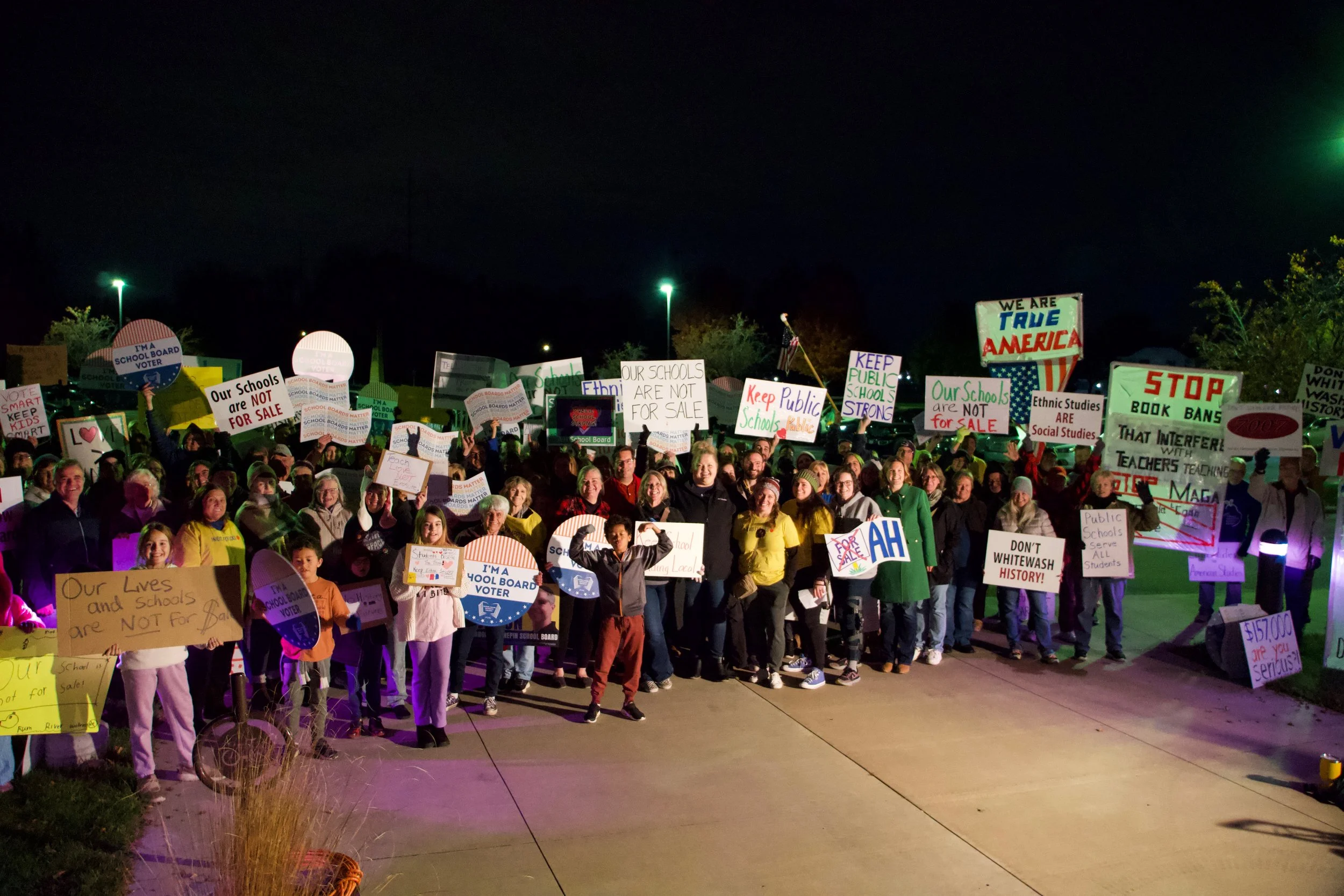A large group of people gathered outdoors at night holding various protest signs supporting public schools and educational issues.