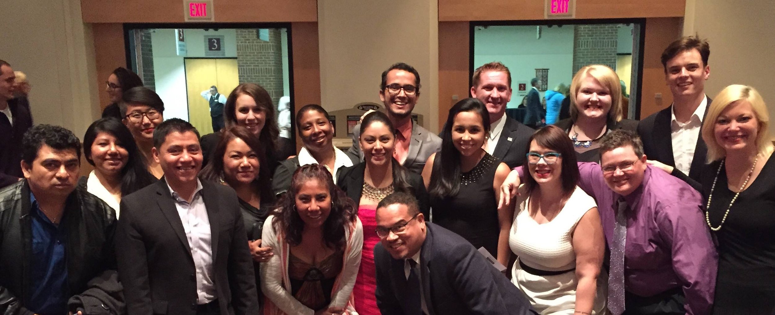 Group of diverse people in formal attire posing for a photo at an indoor event, with two exit signs visible in the background.