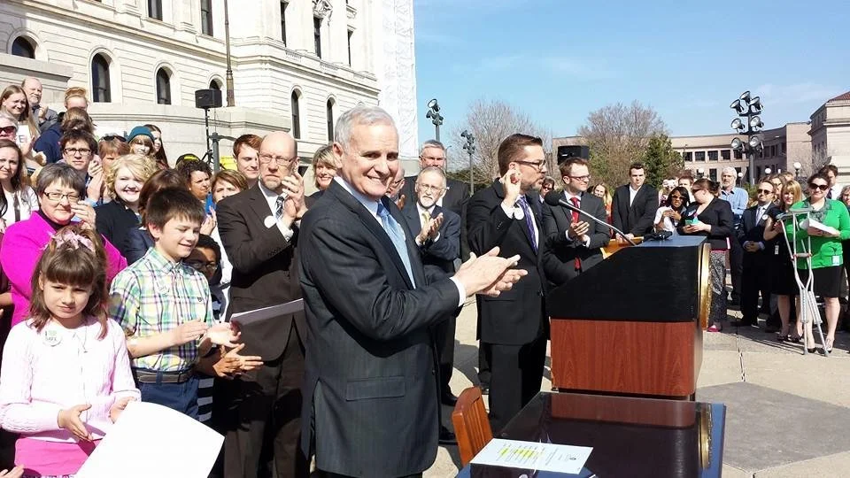A crowd gathered outdoors near a government building, with a man at a podium clapping, and others standing around, some holding papers, watching an event.