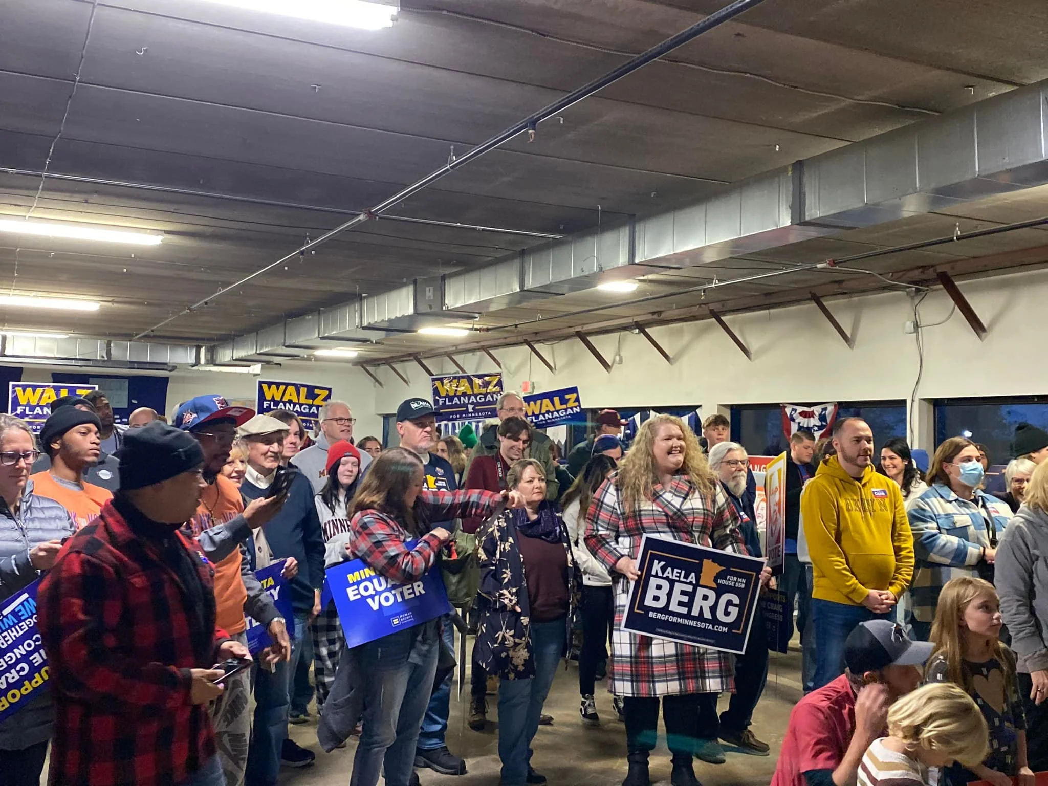 A diverse group of people gathered indoors at a political rally, holding signs supporting a candidate named Kaela Berg. There are campaign signs and banners for Walz and Flanagan in the background.