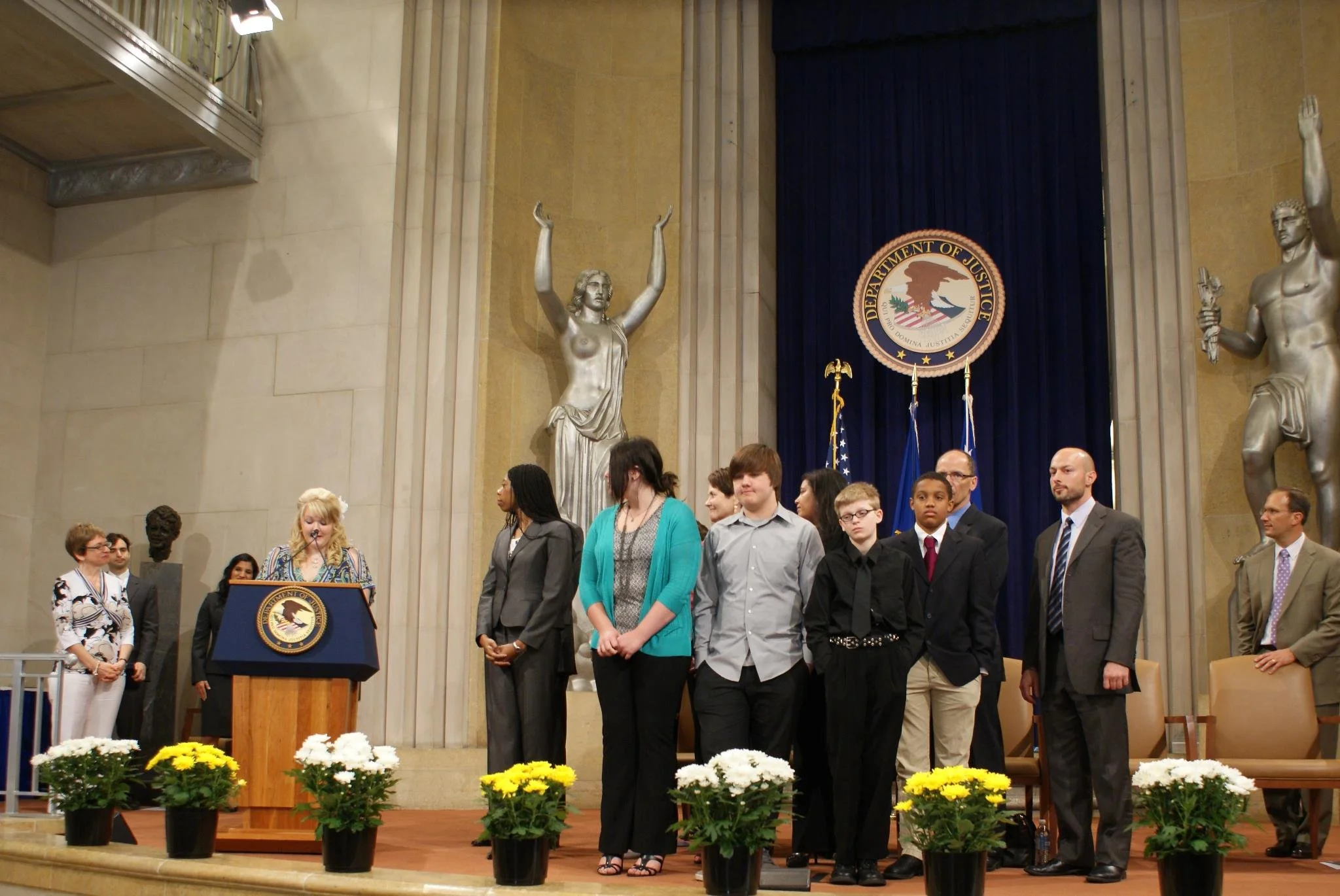 A group of people, including children and adults, standing on a stage in an official government setting, with a woman speaking at a podium bearing the seal of the Department of Justice. The background features large statues and flags.