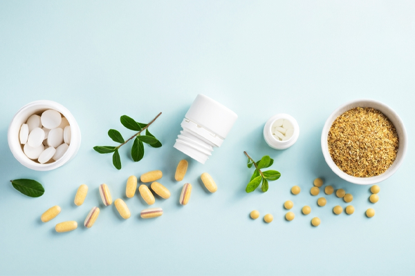 Supplements with white pills, capsules, and powder on a light blue background, alongside green leaves.