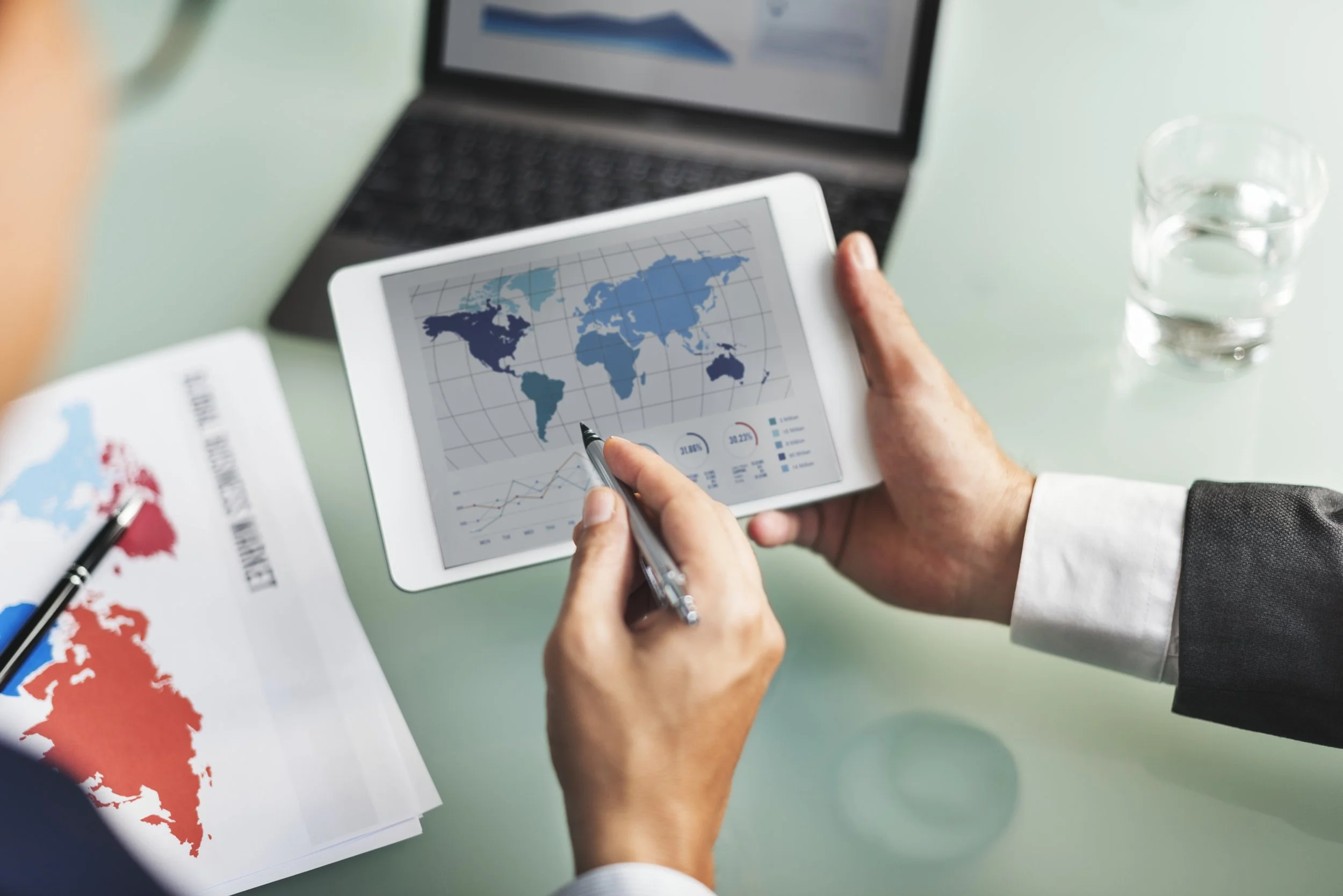 A person in a business suit holding a tablet displaying a world map with data charts in a meeting room. There is a glass of water, a laptop, and a printed world map chart on the table.