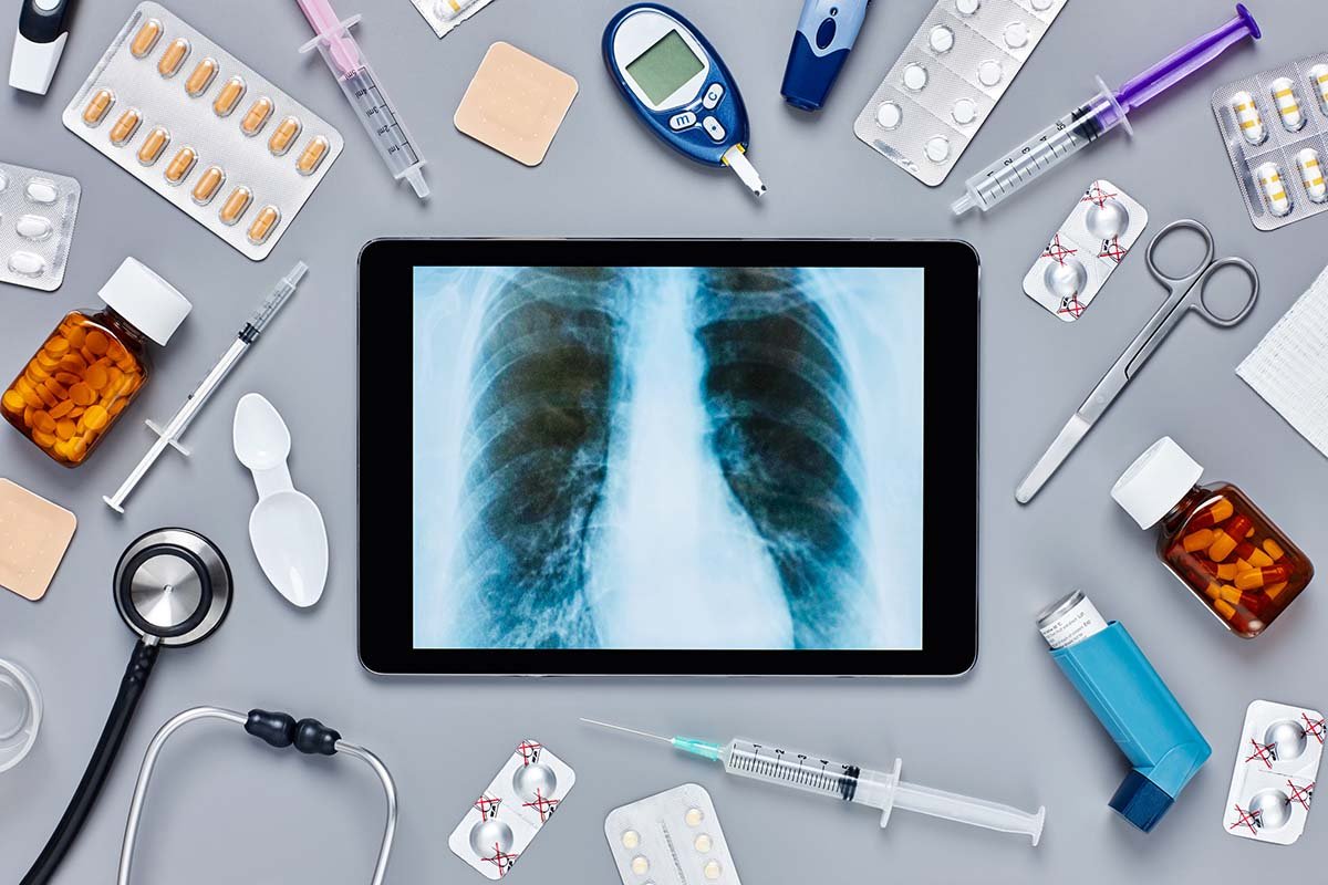 A tablet displaying a chest X-ray image, surrounded by various medical supplies including pills, syringes, a stethoscope, scissors, blister packs, medication bottles, and a glucose meter, on a gray surface.