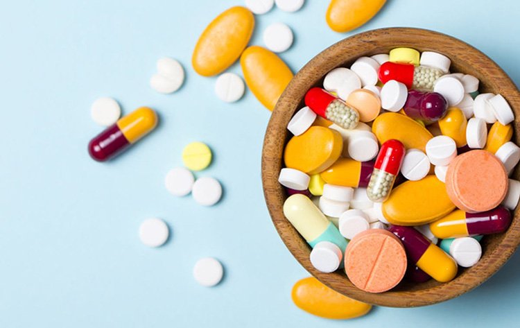 A wooden bowl filled with various colorful pills and capsules, with some pills spilled onto a light blue surface.