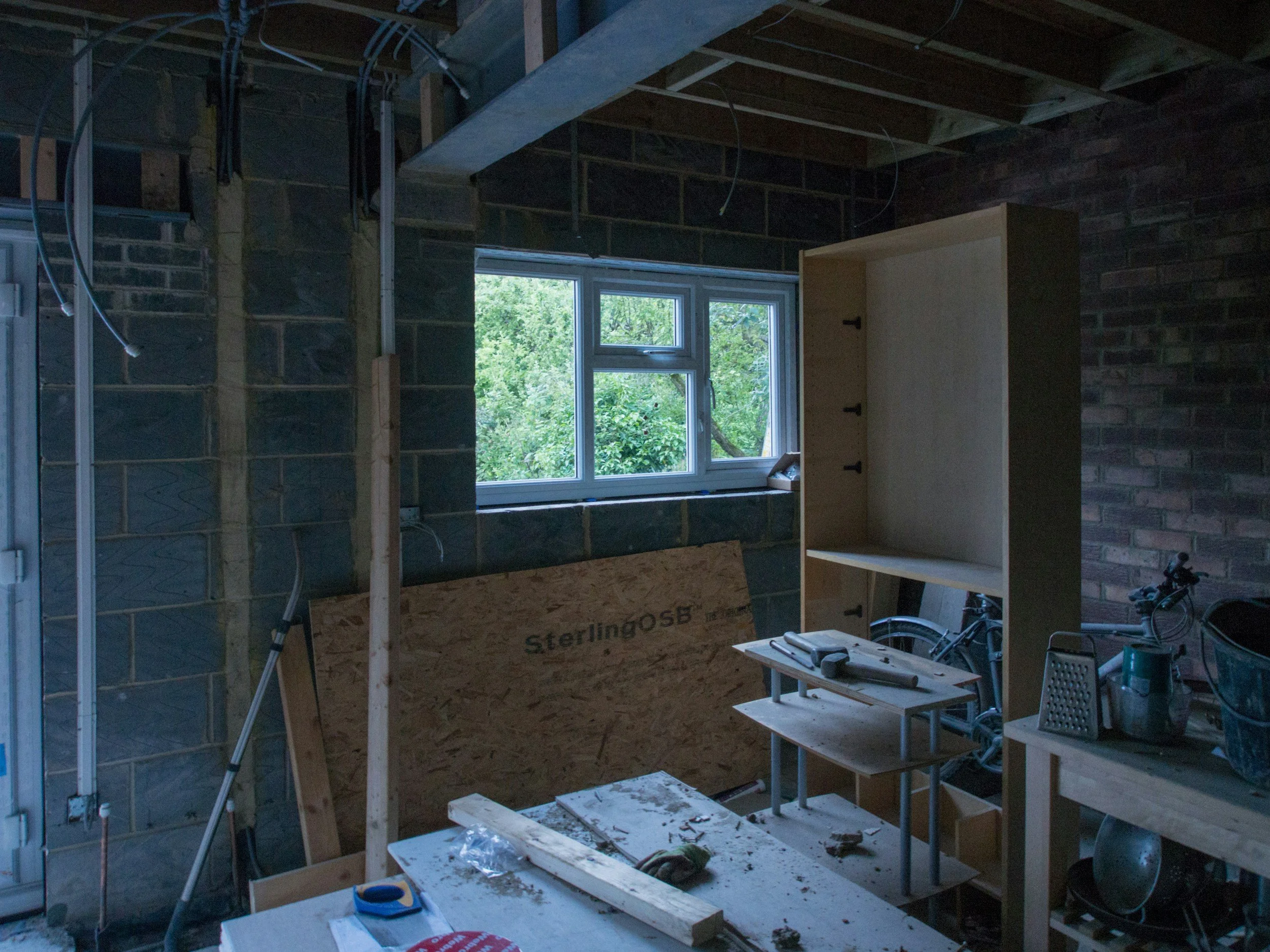 Interior of a room under construction with unfinished brick and concrete walls, a window showing greenery outside, and construction tools and woodwork scattered around.