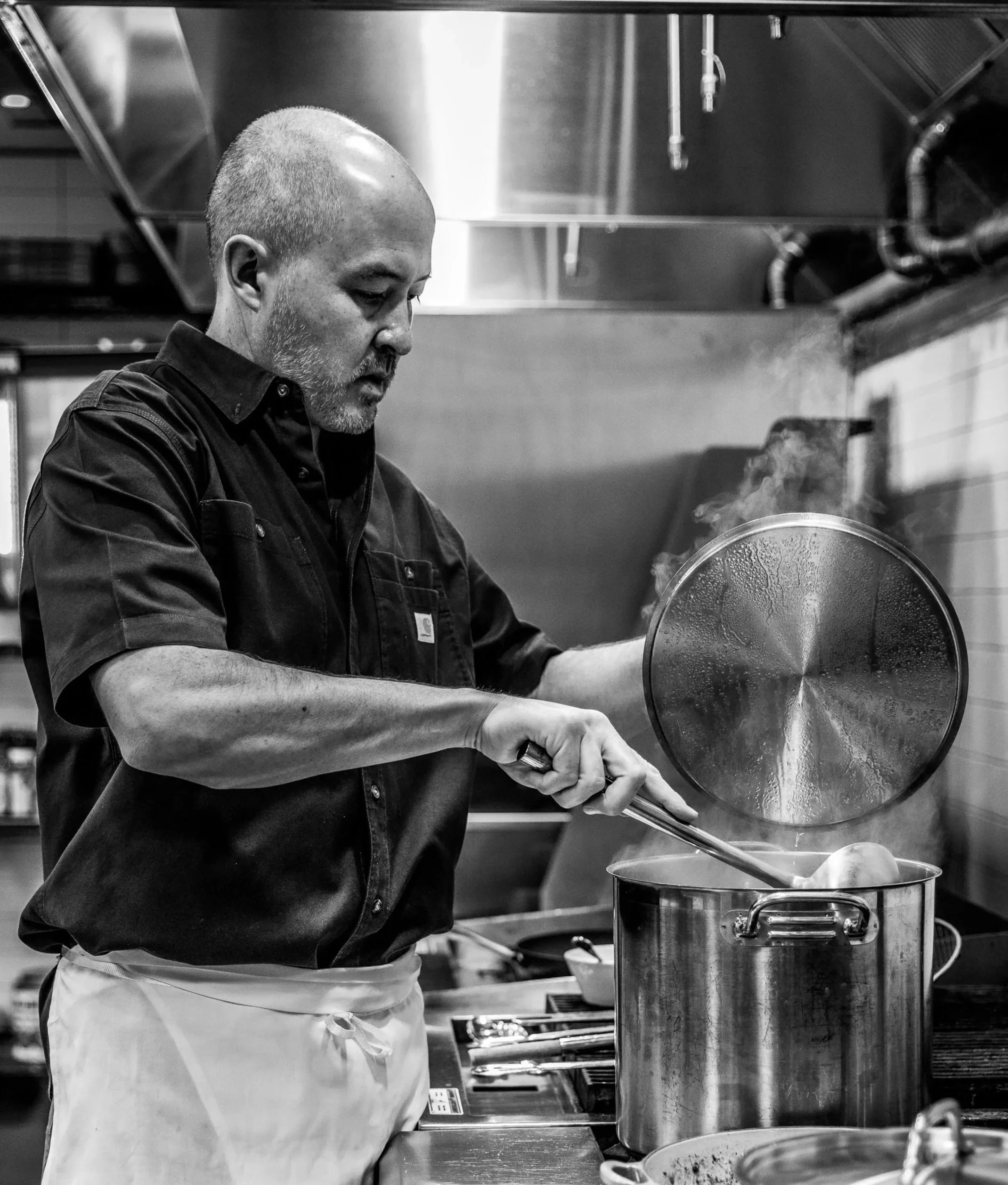 A chef cooking in a commercial kitchen, lifting the lid off a large pot, with steam rising, wearing a black shirt and an apron.