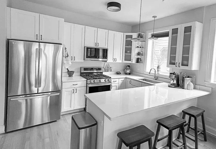 Modern kitchen with white cabinets, stainless steel refrigerator, gas stove, microwave above stove, and a window above the sink. There are three bar stools at the island counter and various kitchen appliances on the counter.