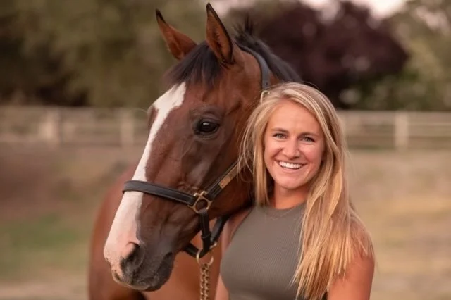 A woman smiling next to a brown horse with a white stripe on its face, outdoors in a fenced area.