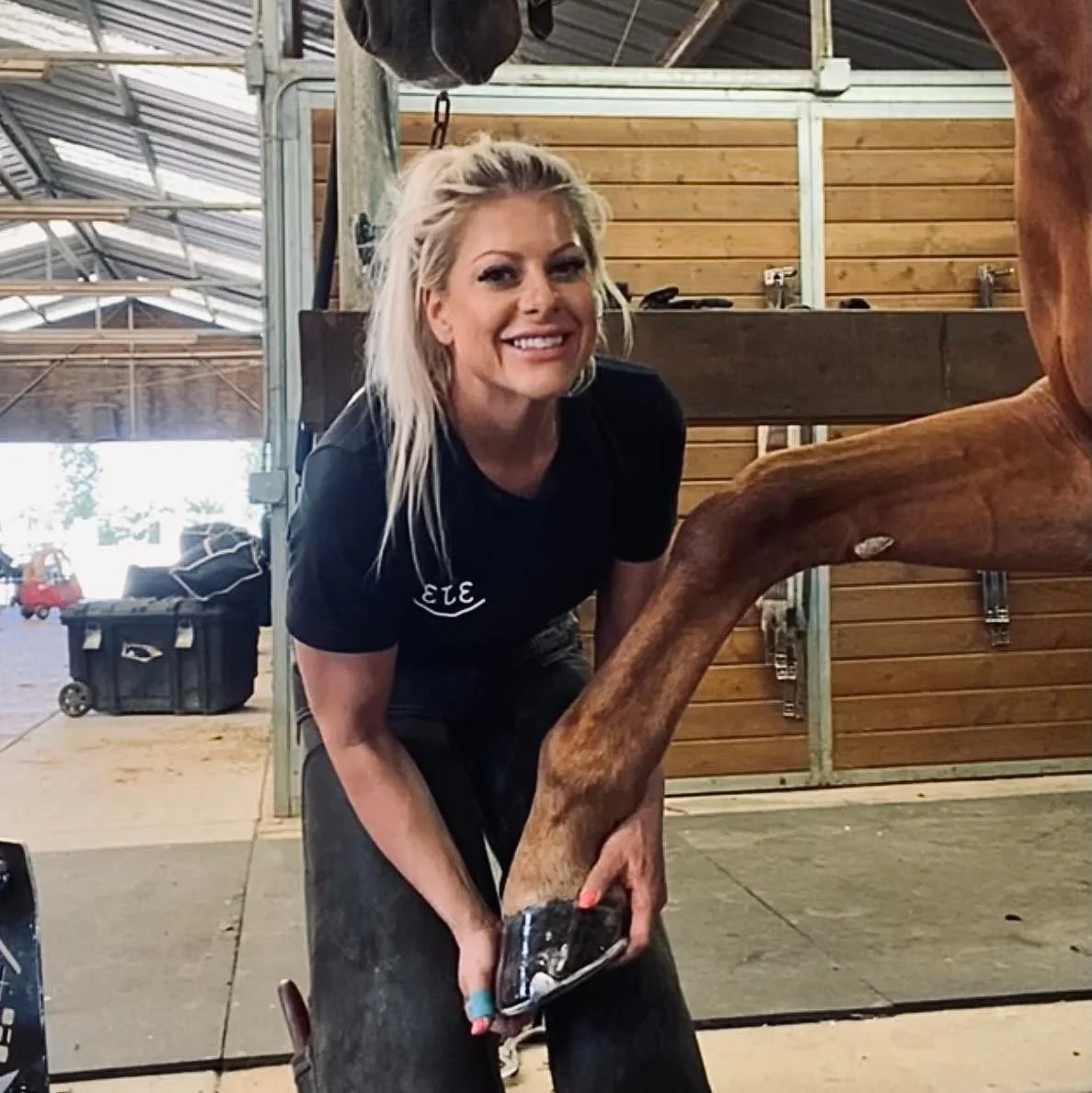 Woman smiling and holding the leg of a horse in a stable with wooden walls and metal roof construction.