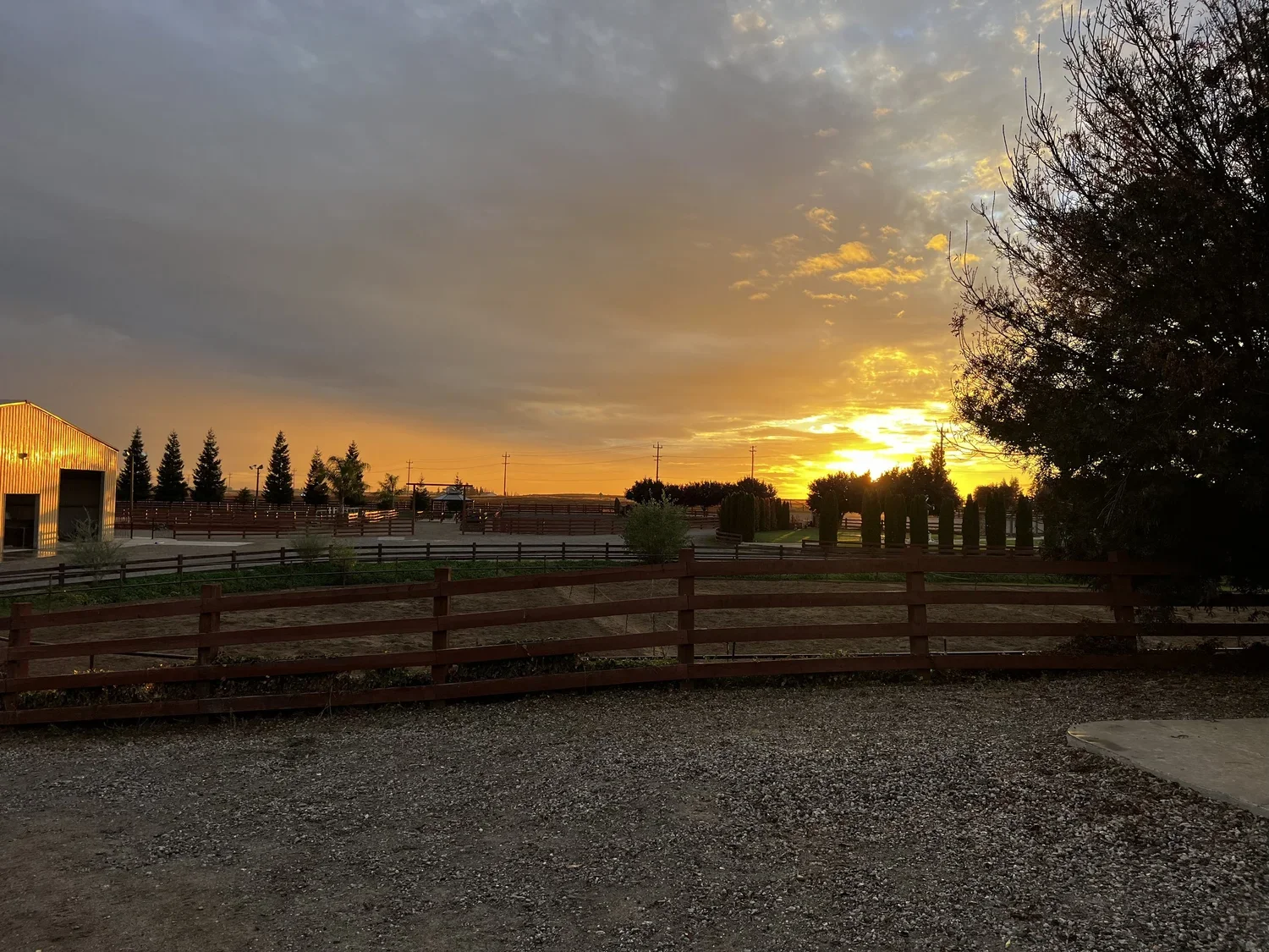 Sunset over a fenced rural area with trees, a building on the left, and a gravel ground in the foreground.