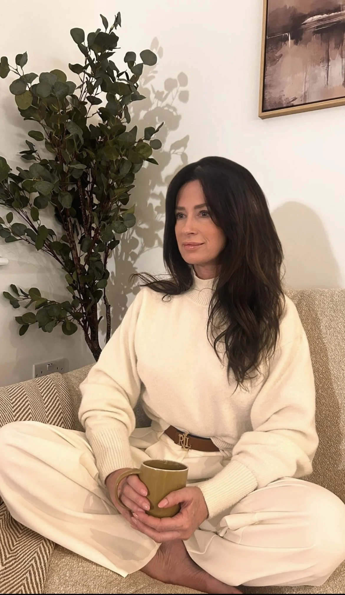 Woman sitting cross-legged on a beige couch, holding a green mug, wearing a cream-colored sweater and white pants, with a large green potted plant and a framed painting on the wall behind her.