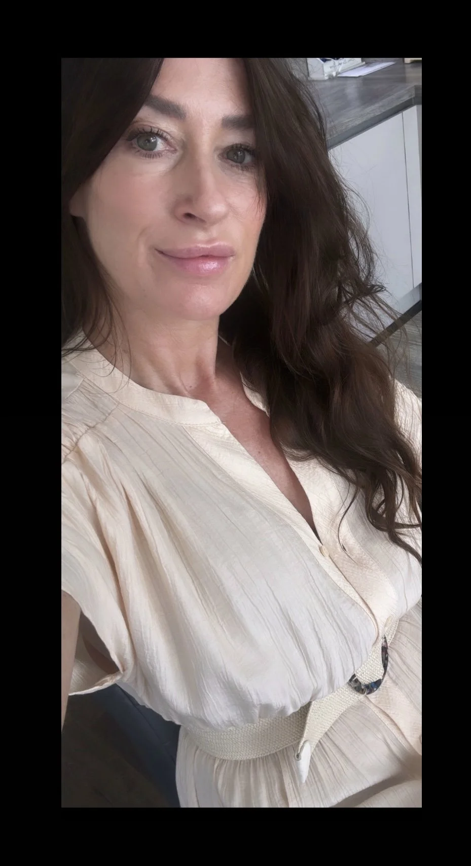 Close-up of a woman with long brown wavy hair, light skin, wearing a cream-colored blouse and sitting in an indoor setting.