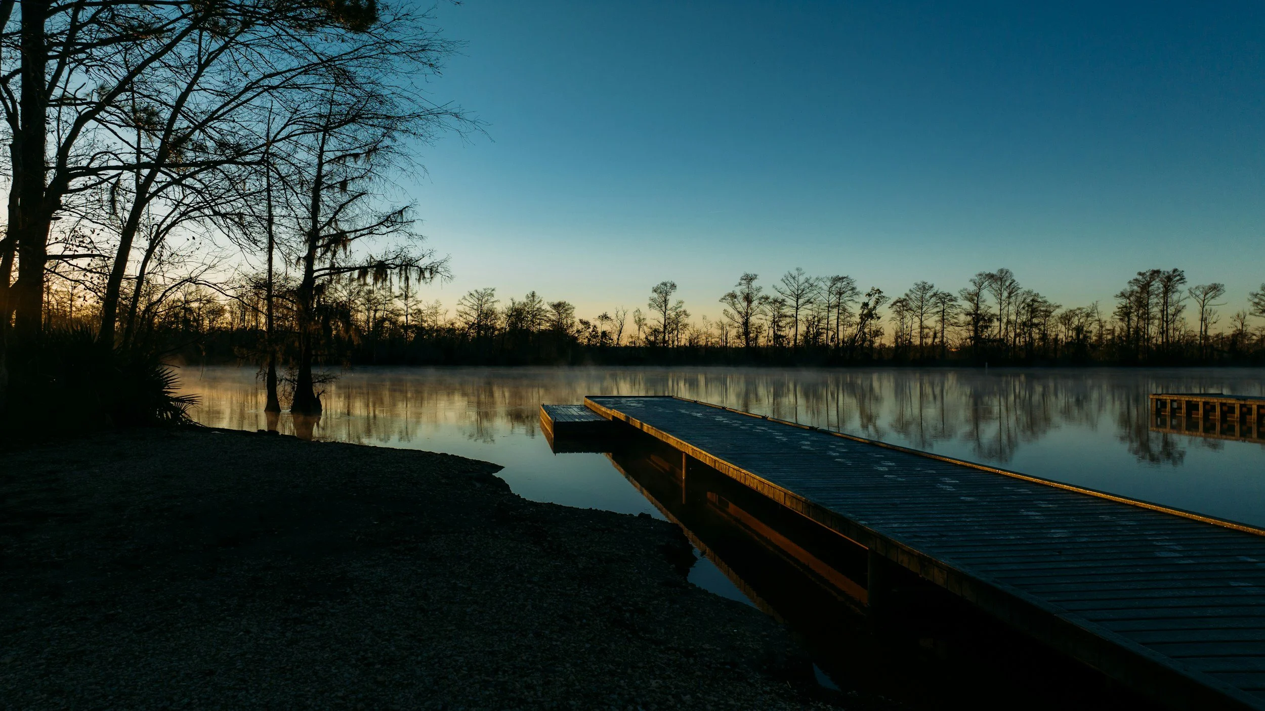 A peaceful lakeside scene during sunrise or sunset with a wooden dock extending into calm water, surrounded by leafless trees and reflections in the water.