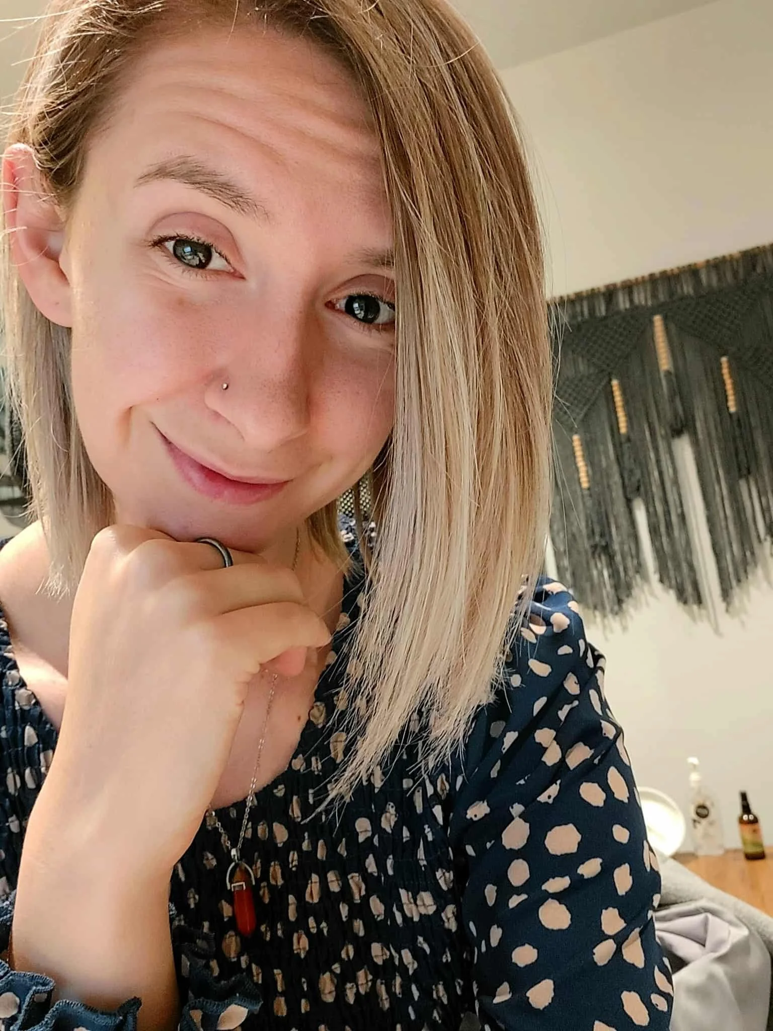 A young woman with blonde hair and a nose piercing, smiling softly and resting her chin on her hand, wearing a dark blouse with a beige polka dot pattern. In the background, there is a decorative wall hanging and some bottles on a table.