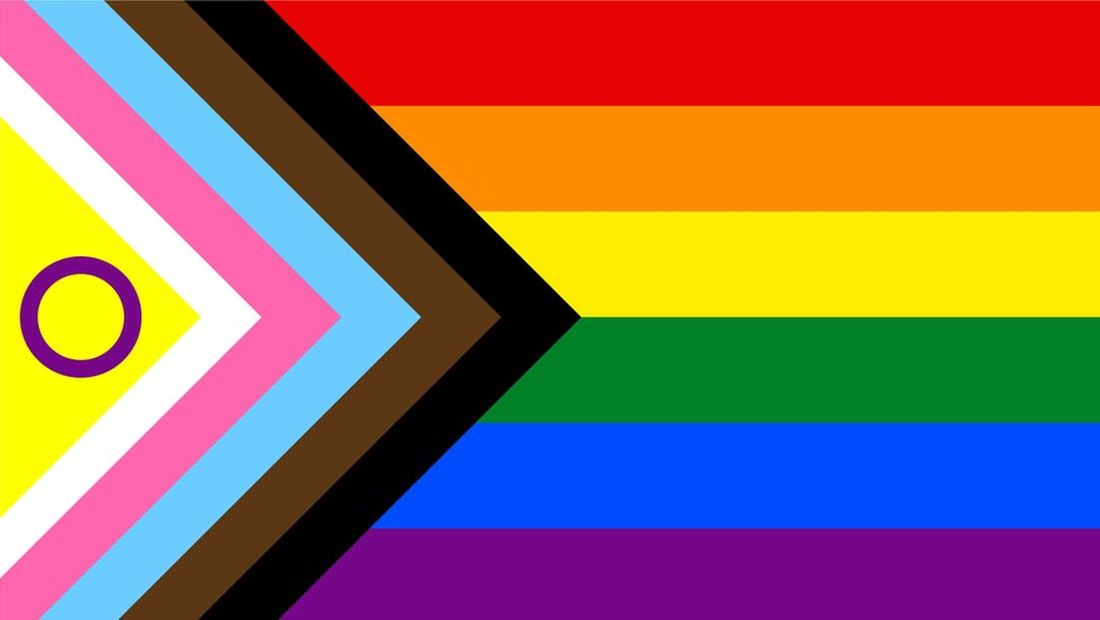 A Pride flag with rainbow stripes and a black and brown stripe, a pink, white, and blue stripe, and a purple circle on a yellow triangle.
