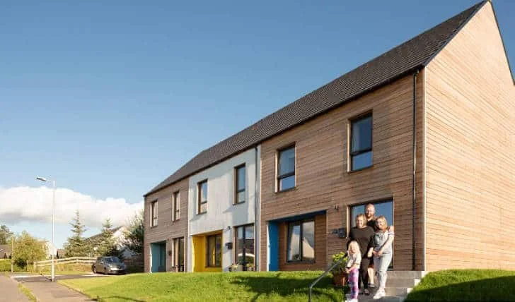 Family standing outside a modern townhouse with wooden and white facades, a yellow door, and a small front yard.