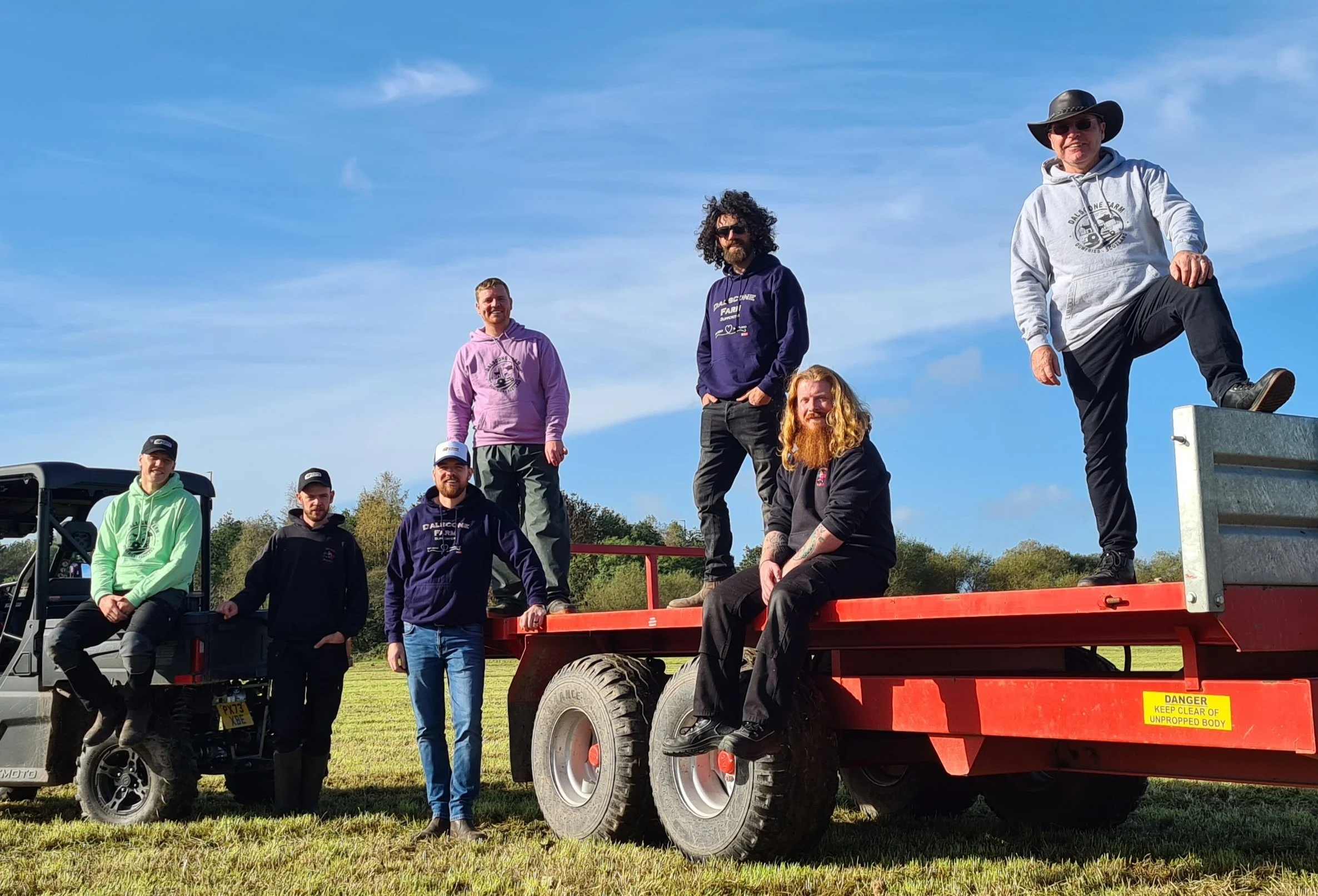Group of men posing on and around a red flatbed trailer, with some sitting and others standing, outdoors on grass under a blue sky.