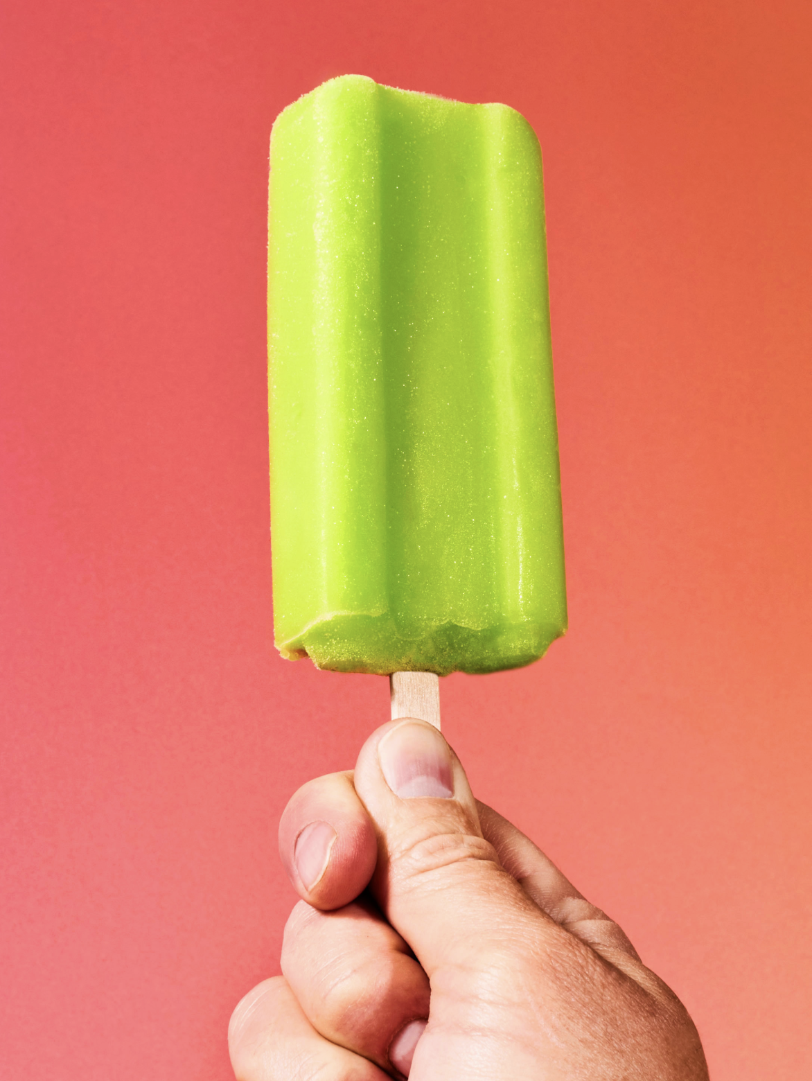A hand holding a bright green popsicle against a coral pink background.