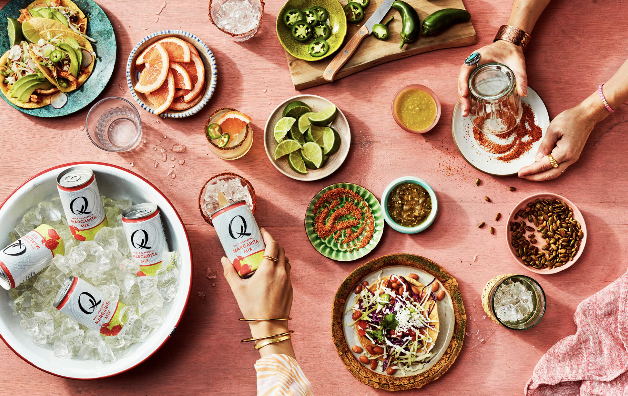 Top-down view of a colorful Mexican meal spread including tacos with avocado and lime, assorted salsas, lime wedges, jalapeños, margarita drinks, and a bowl of beer on a pink table.