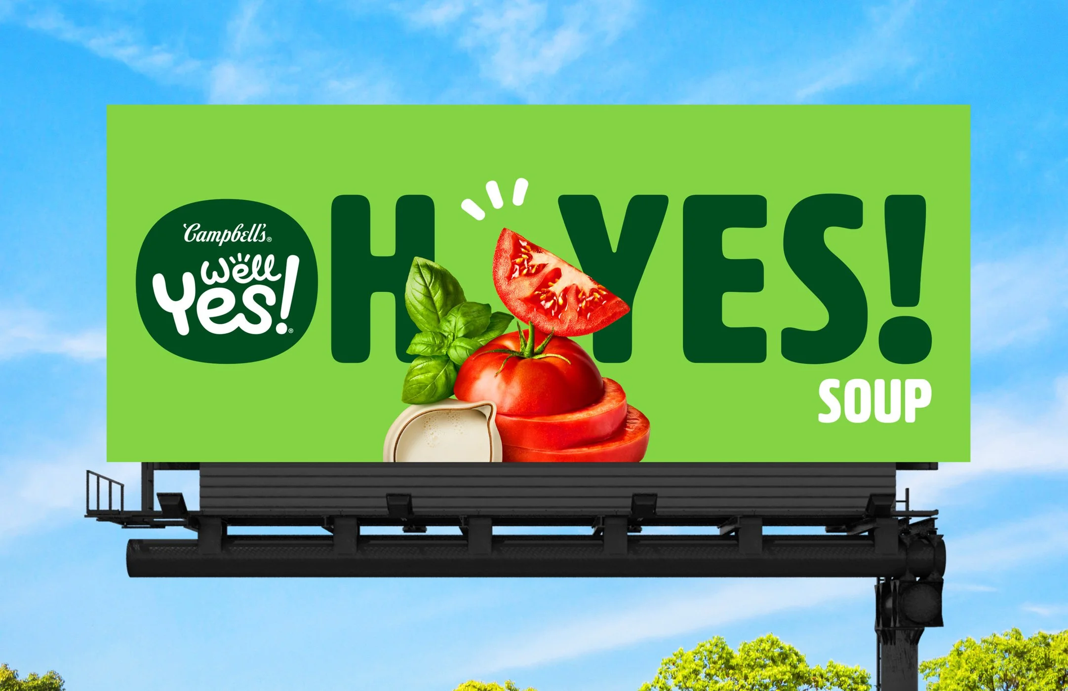 Billboard advertising Campbell's Well Yes! Hummus Soup with images of tomato, basil, and mozzarella cheese on a green background against a blue sky.