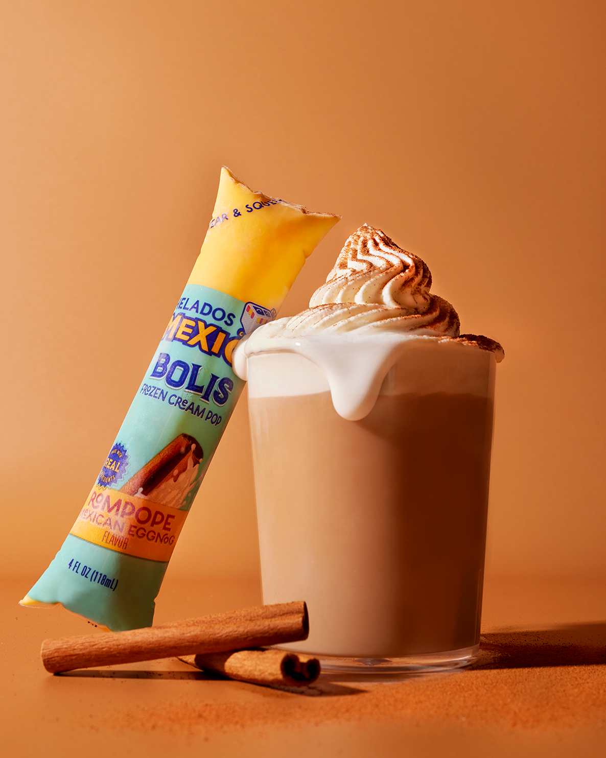 A cup of Mexican eggnog ice cream topped with whipped cream and sprinkled with cinnamon, with a cinnamon stick beside it, and an ice cream popsicle in colorful packaging leaning against the cup.
