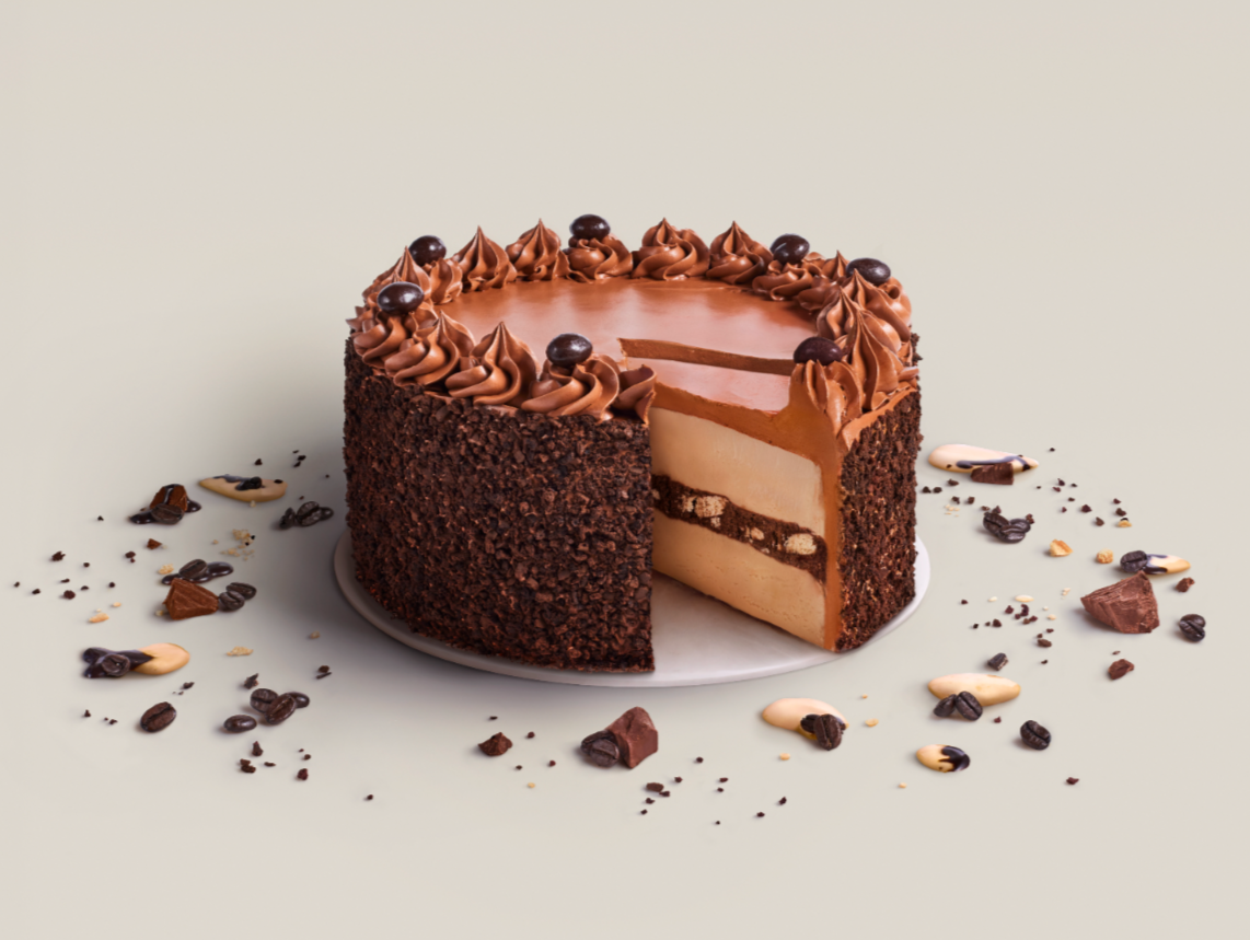 Chocolate cake with a layer of light-colored cream filling, decorated with swirls of chocolate frosting and chocolate-covered toppings, surrounded by scattered chocolate pieces and coffee beans.