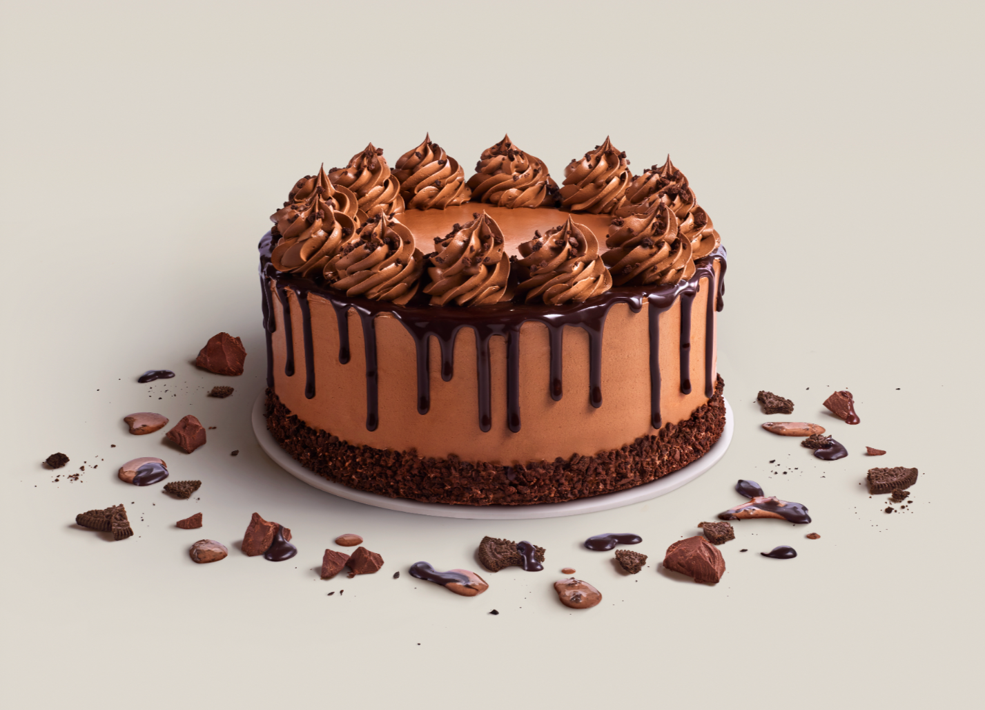 Chocolate cake with chocolate frosting, chocolate drizzle, and whipped frosting swirls on top, surrounded by scattered chocolate pieces and crumbs on a light-colored surface.