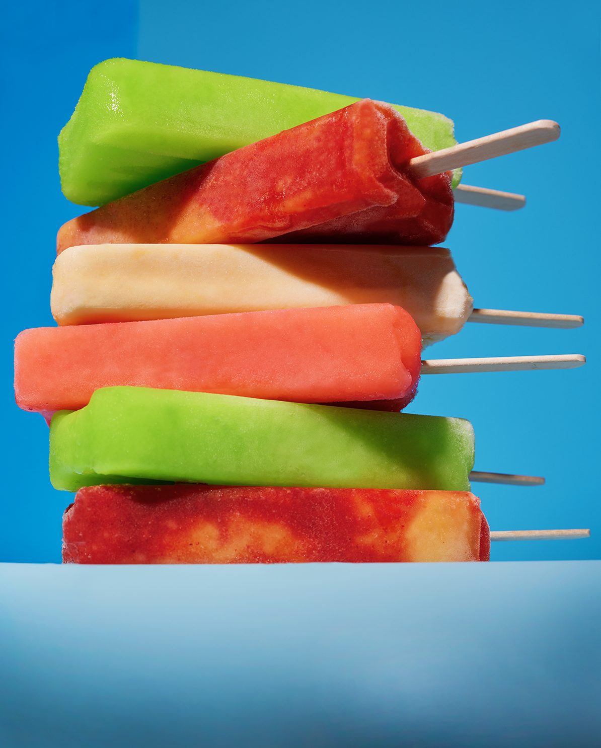 Stack of colorful frozen popsicles on a light blue surface with a bright blue background.