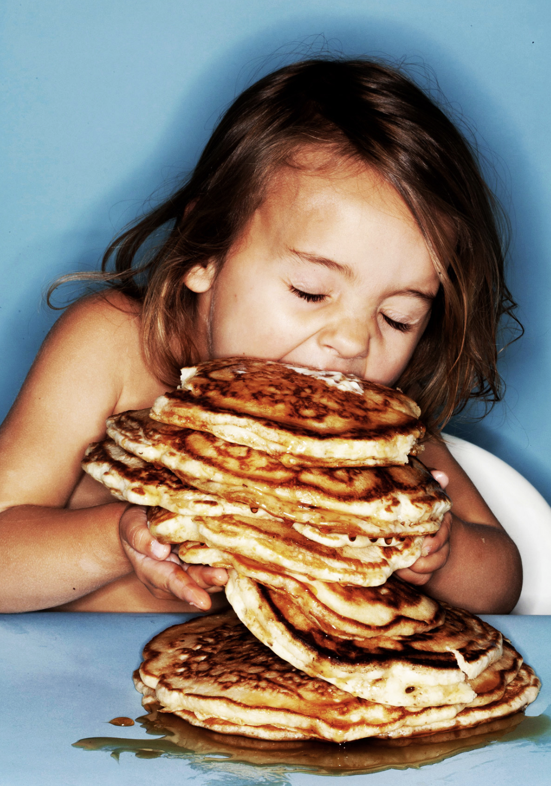 A young girl with light skin and brown hair is holding and about to eat a large stack of pancakes with syrup on a blue background.