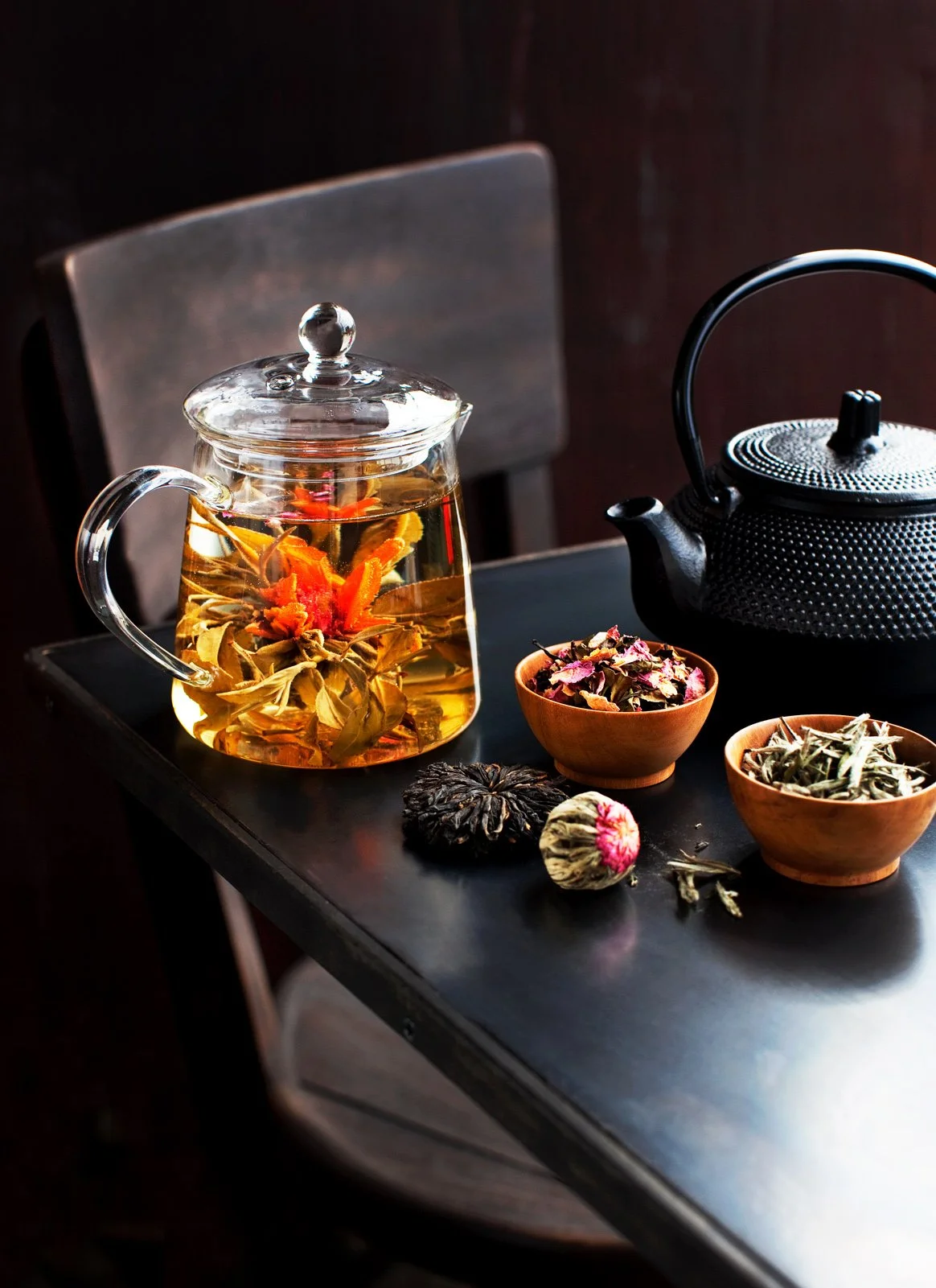 A glass teapot filled with herbal tea, a black cast iron teapot, and two wooden bowls containing dried herbs and flower petals on a black table.