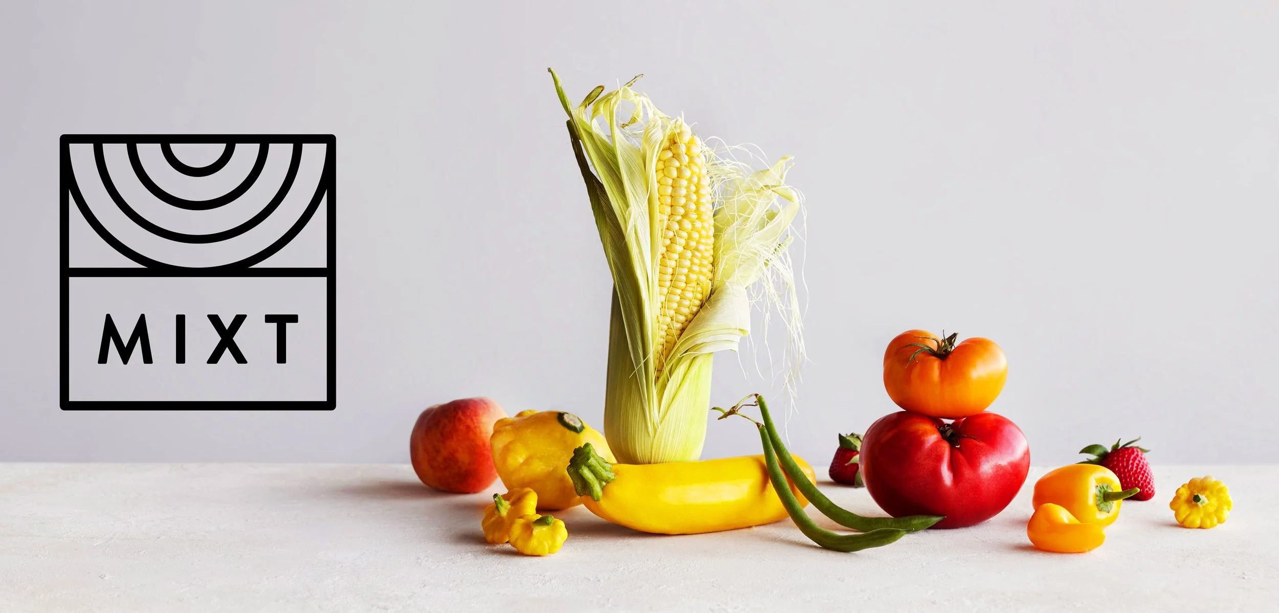 A variety of fresh vegetables and fruits including an ear of corn, a yellow bell pepper, a banana, two tomatoes, a strawberry, an orange bell pepper, and small yellow peppers arranged on a white surface against a light gray background. A black logo w