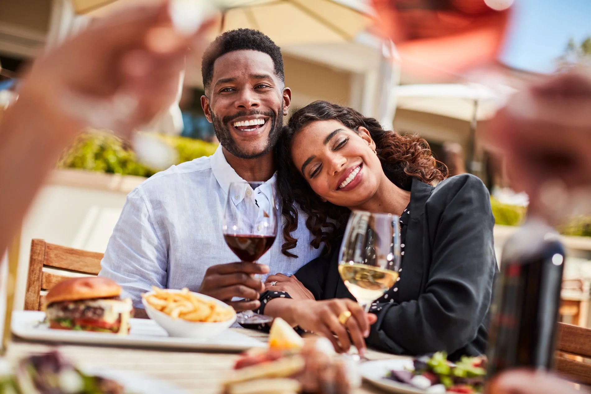 A smiling man and woman enjoying drinks and food at an outdoor dining table, with other people and patio umbrellas in the background.
