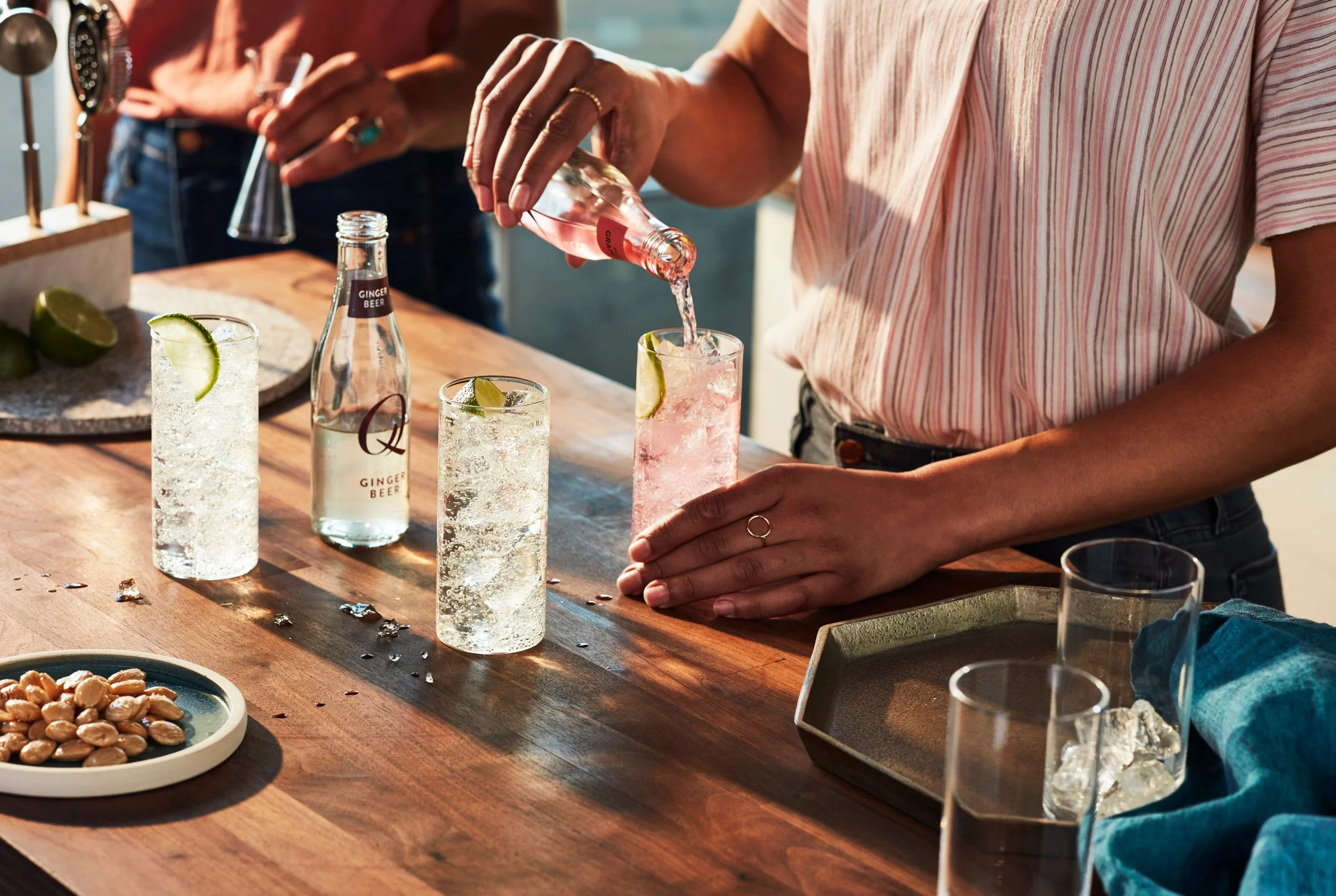 Person pouring pink drink into a glass with lime wedge at a bar, with other drinks and ingredients on the wooden counter.