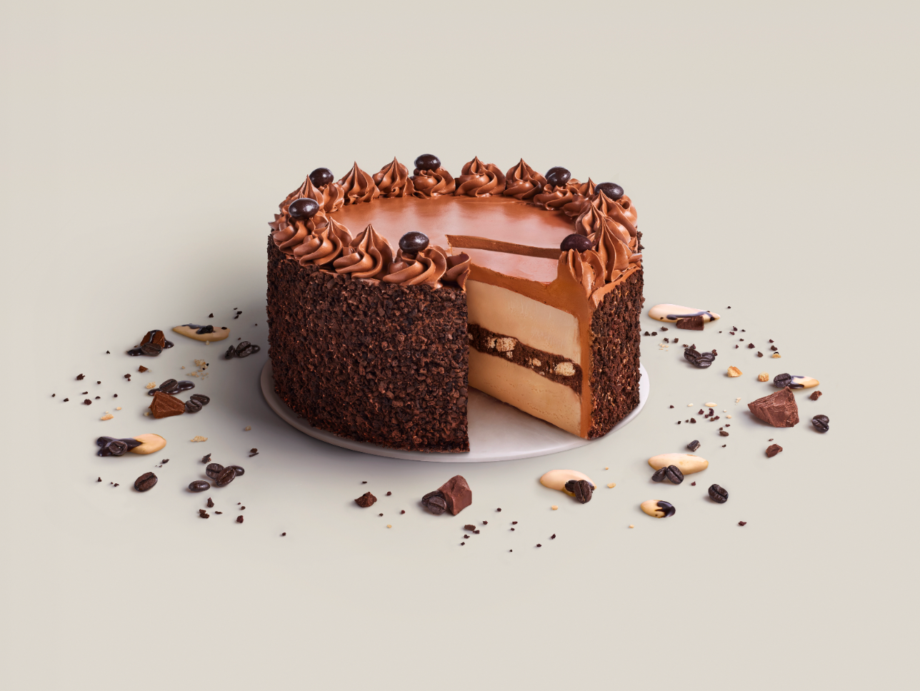 Chocolate cake with a layer of light-colored cream filling, decorated with swirls of chocolate frosting and chocolate-covered toppings, surrounded by scattered chocolate pieces and coffee beans.