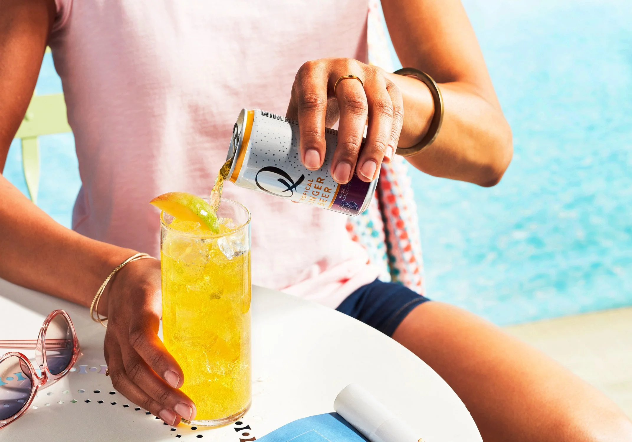 A person is pouring a can of hard seltzer into a tall glass of ice and lemon slices, sitting outdoors near a body of water.