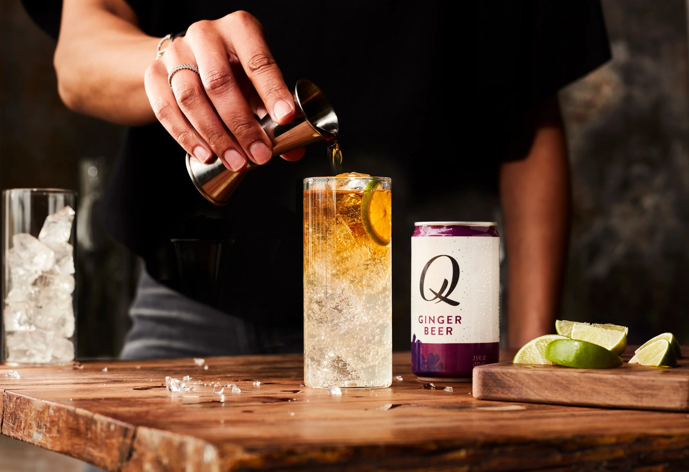 Person pouring a ginger beer cocktail into a tall glass with lime slices on it, on a wooden table with lime wedges and a can of ginger beer nearby.