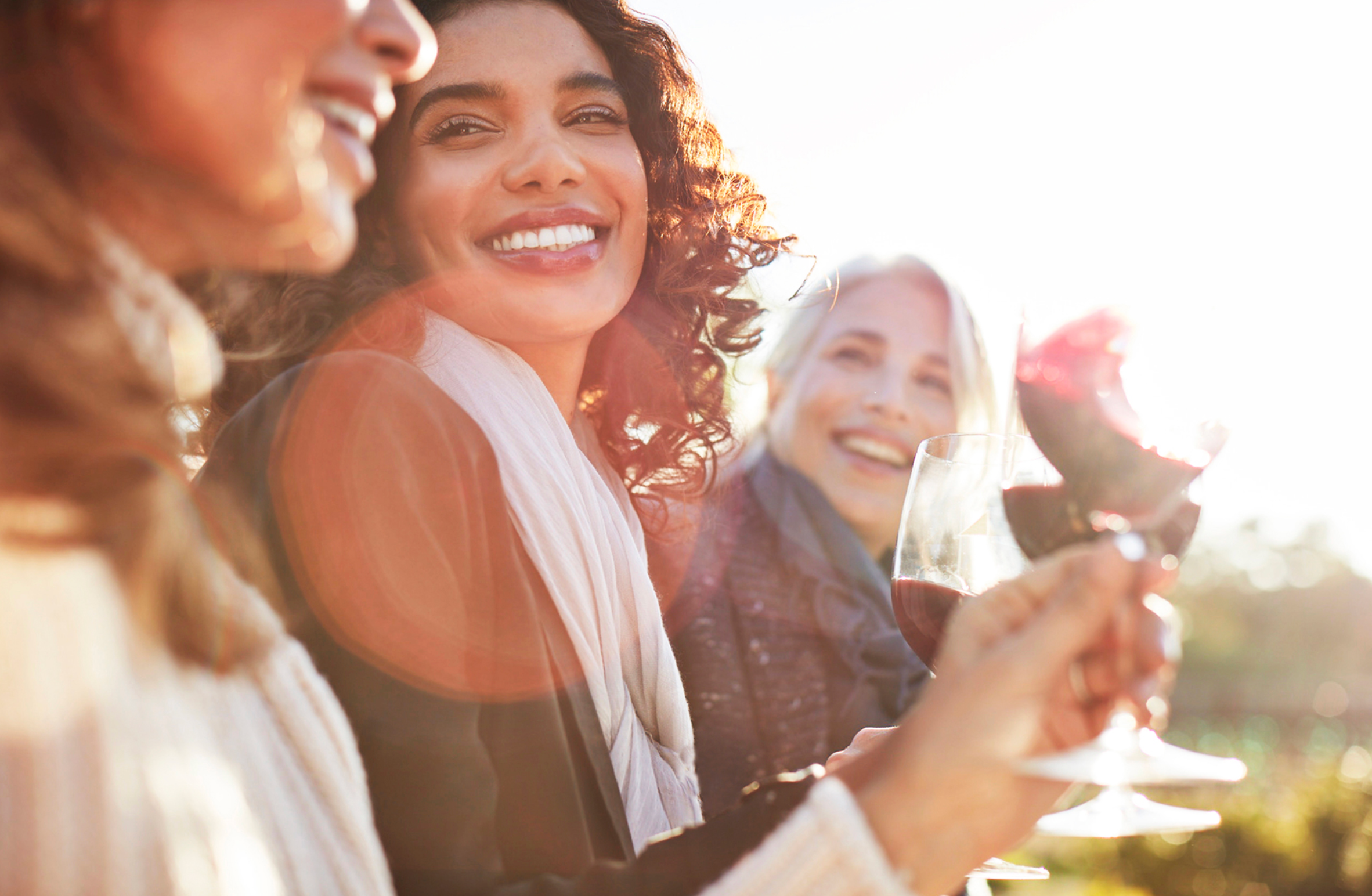 Three women smiling and enjoying red wine outdoors in the sunlight.