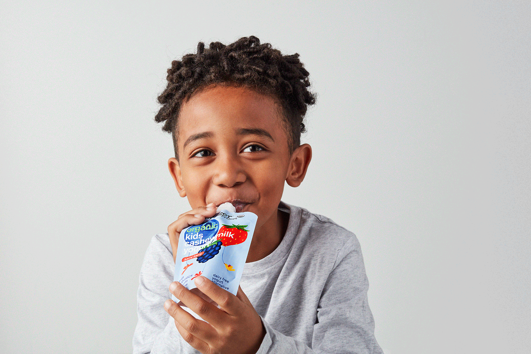 Young boy with curly hair drinking organic kids yogurt, smiling and looking at the camera, against a plain light gray background.