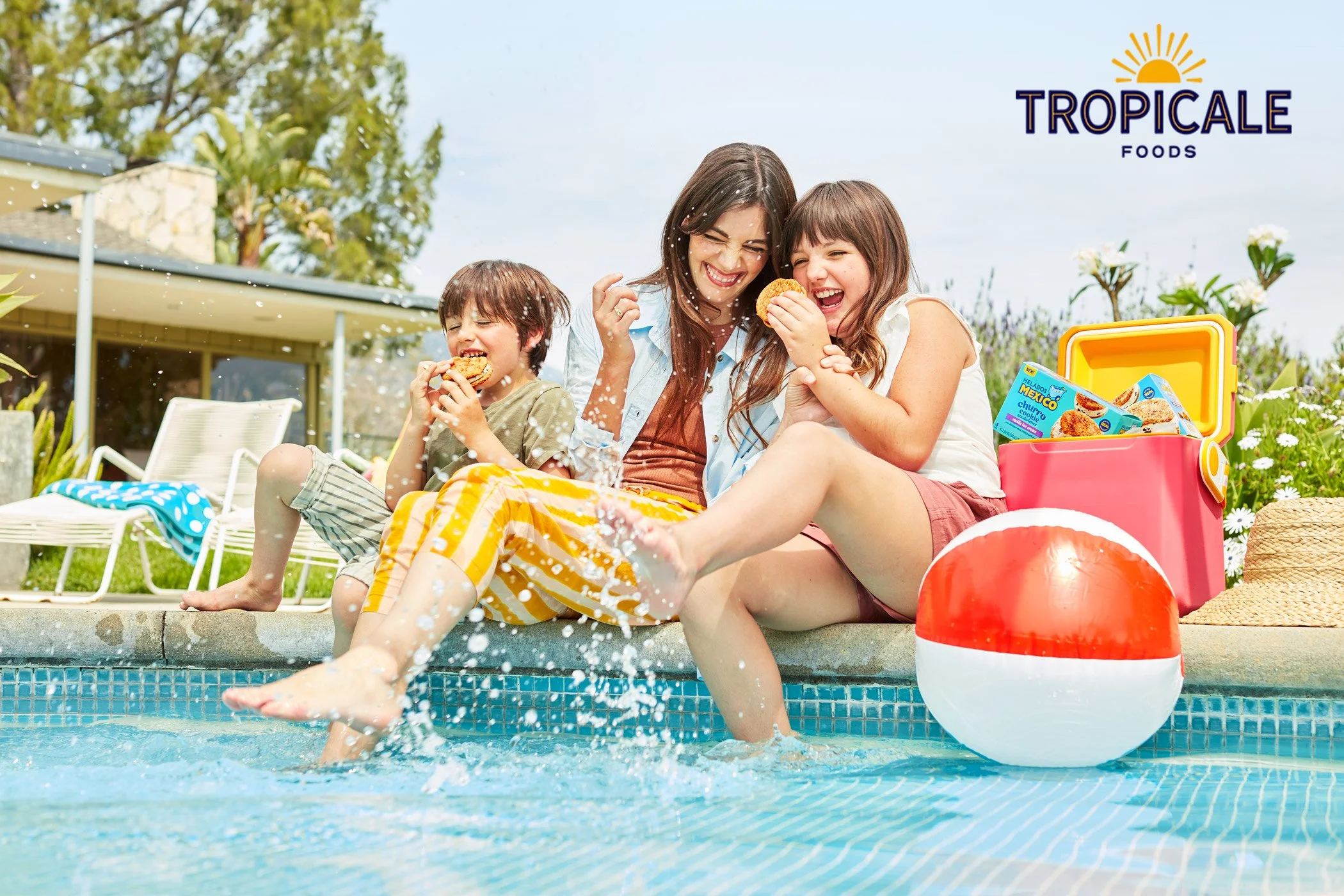 A woman and two children sitting by a swimming pool, eating cookies, with a red and white beach ball, cooler, and tropical plants in the background.