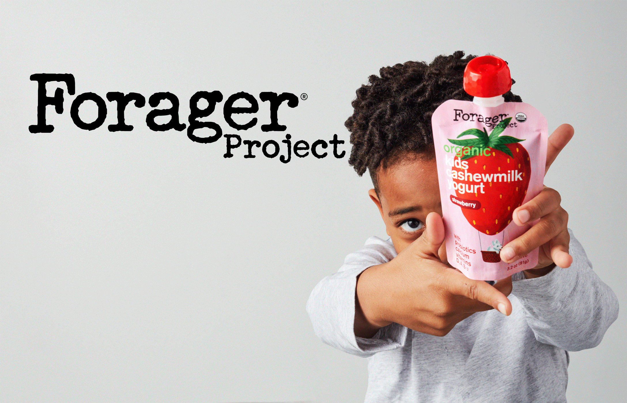 A young child with curly hair holding a pink pouch of strawberries flavored CashewMilk yogurt close to their face, with only their eyes and hands visible, in front of a plain background with the Forager Project logo.