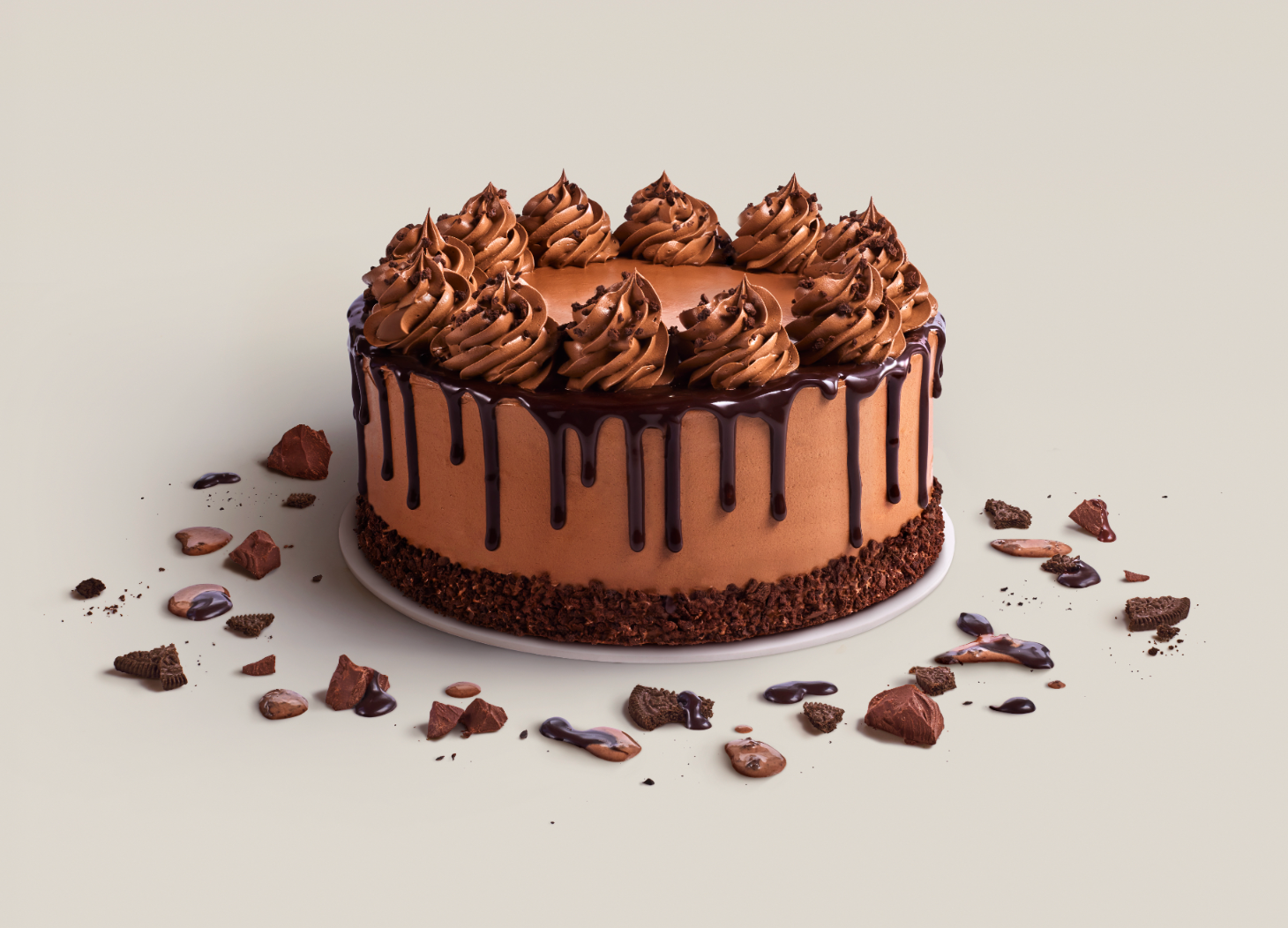 Chocolate cake with chocolate frosting, chocolate drizzle, and whipped frosting swirls on top, surrounded by scattered chocolate pieces and crumbs on a light-colored surface.
