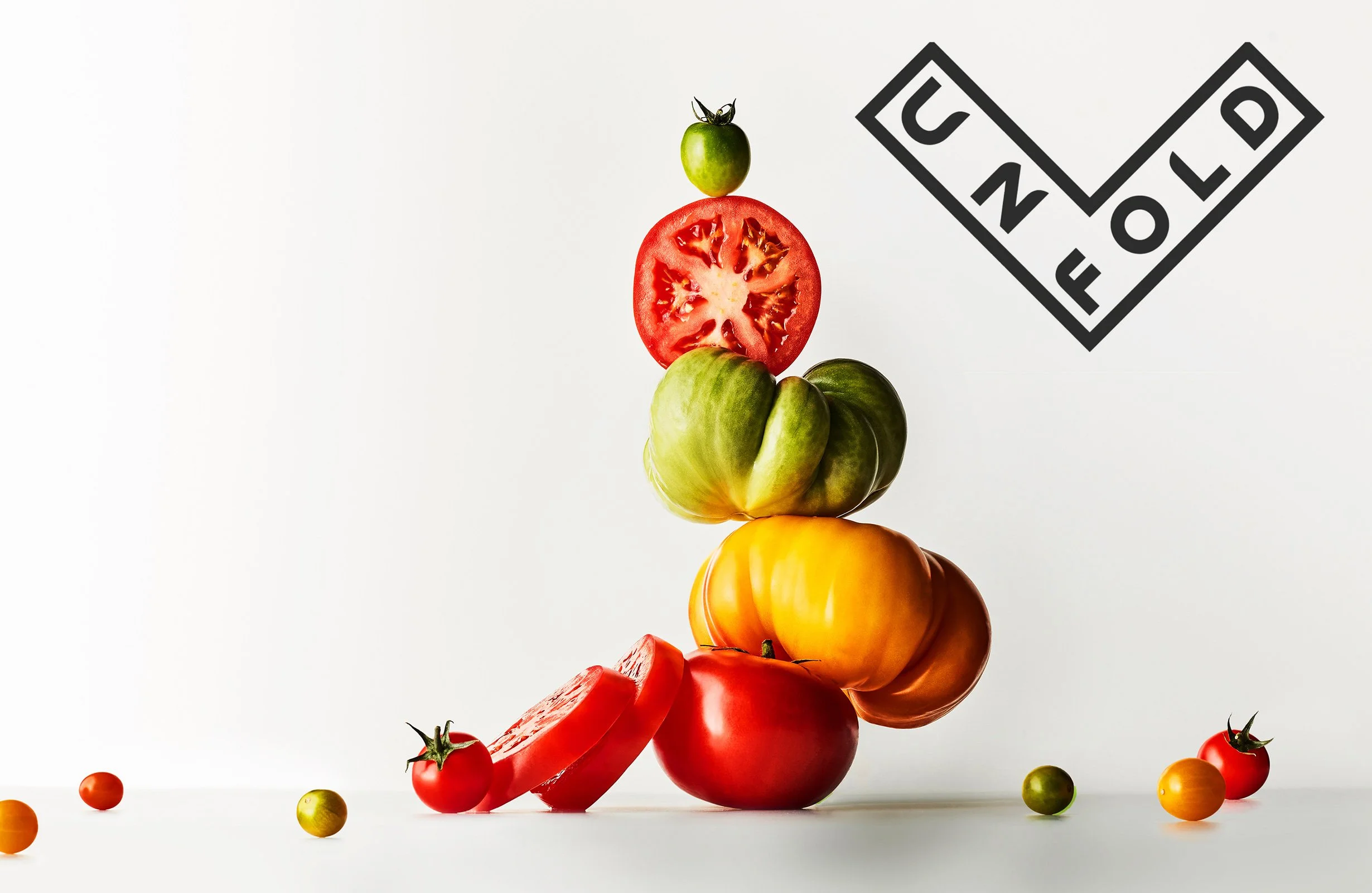 A stack of various colorful tomatoes, including red, green, yellow, and orange, arranged vertically against a white background with a black and white 'C NL D' film clapperboard in the upper right corner.